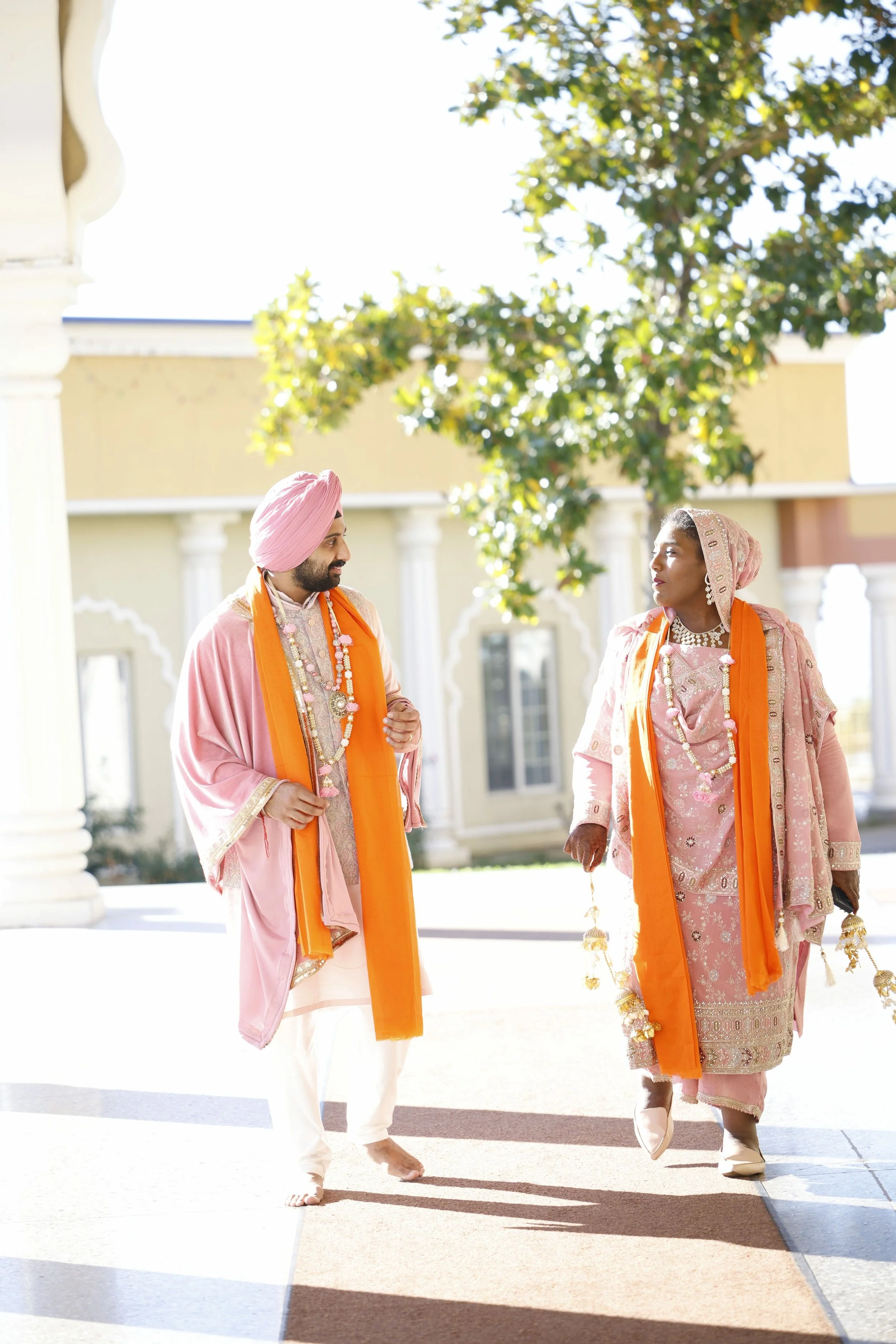 A man and a woman dressed in traditional Indian wedding attire, walking outdoors on a sunny day with clear skies, surrounded by a building with columns and green trees in the background.