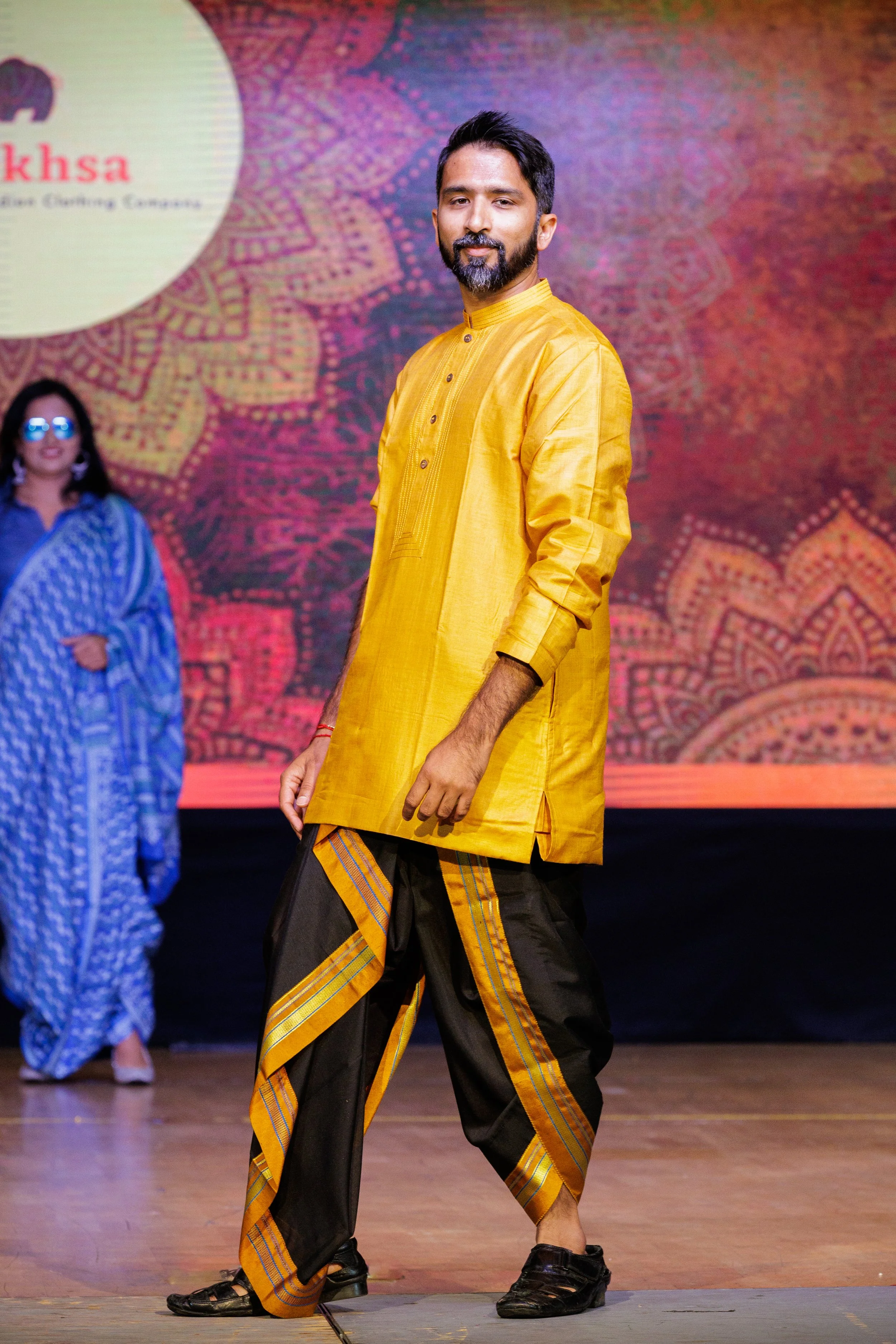A man wearing an orange kurta and black and orange dhoti pants standing on a stage, with a woman dressed in blue saree in the background. There is a decorative backdrop with a logo and text.