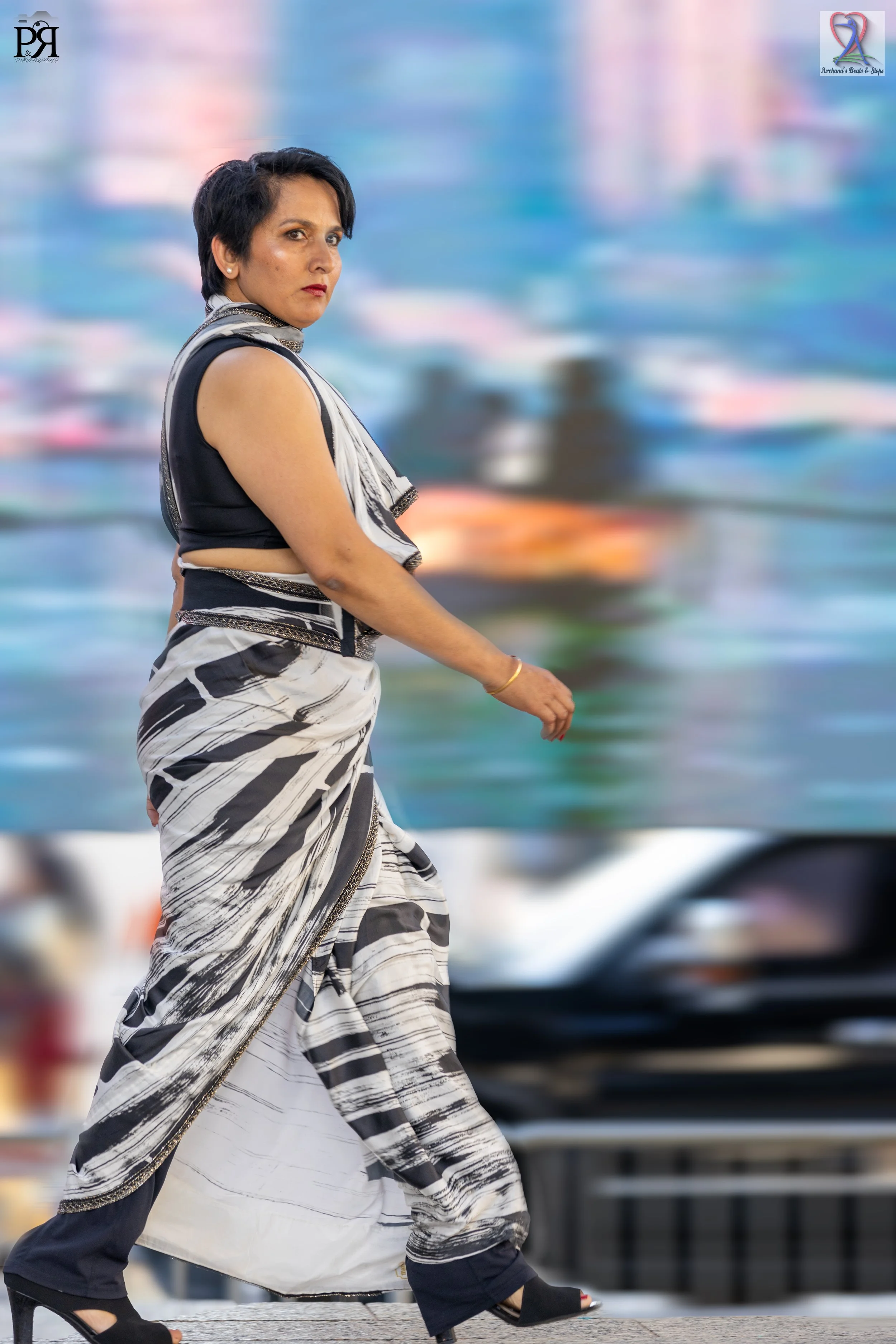 A woman walking outdoors on a city street, wearing a black and white patterned sari and black high-heeled shoes, with blurred colorful cityscape background.