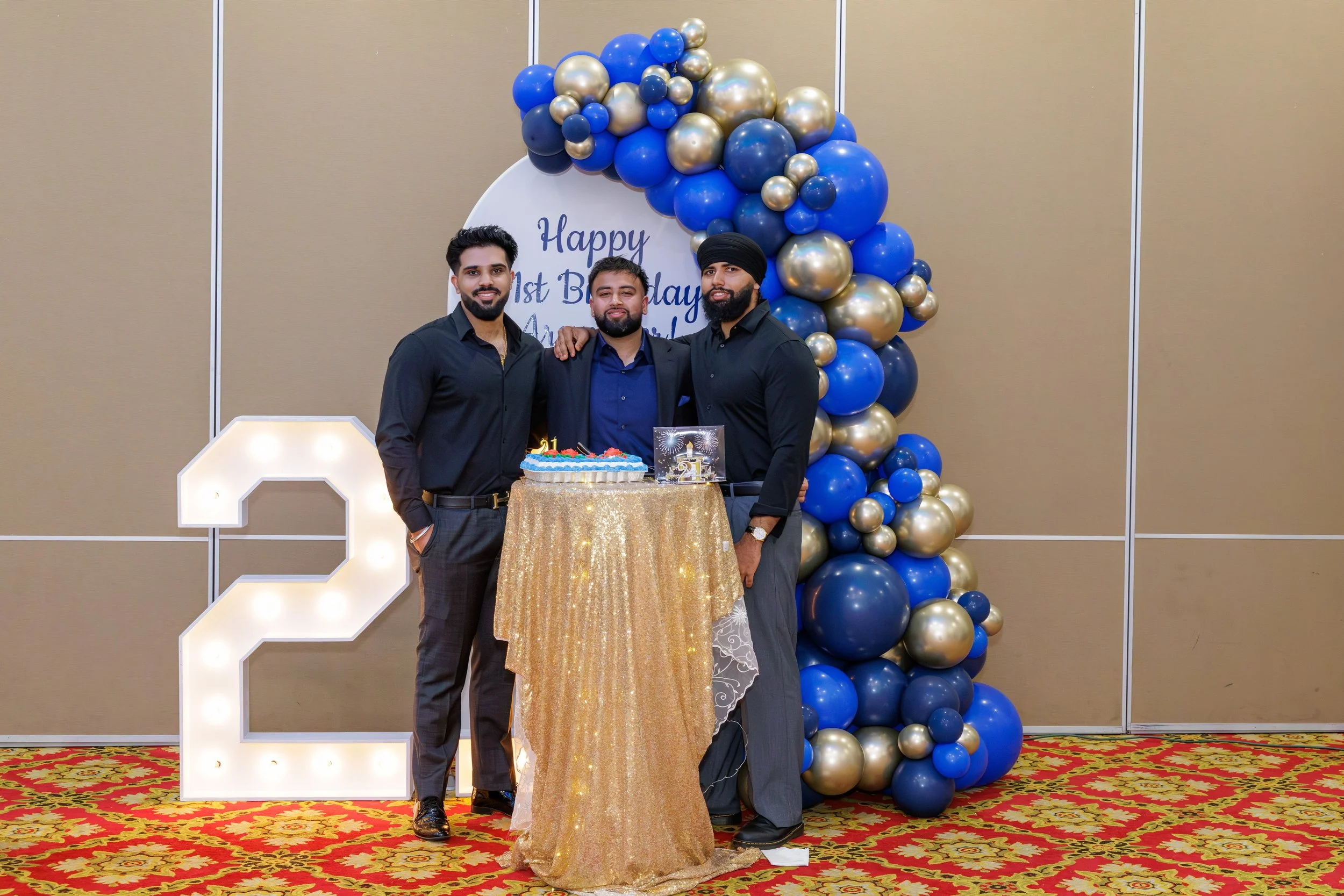 Three men standing together at a birthday celebration with a decorated backdrop, a large illuminated number '2', and a table with a birthday cake.