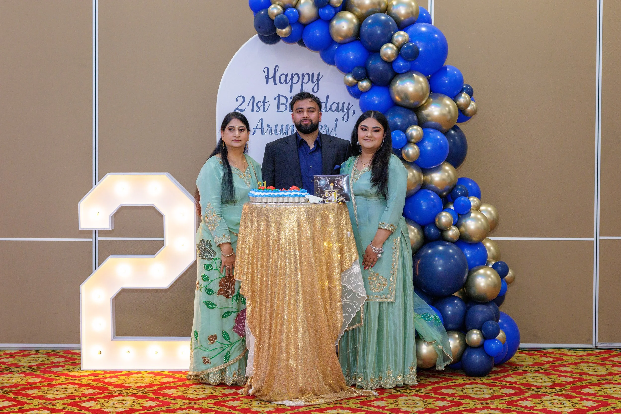 Three people celebrating a 21st birthday standing behind a round table with a cake, in front of a decorated backdrop with balloons and a lit number 2. The backdrop has a banner that reads 'Happy 21st Birthday, Aruna!'