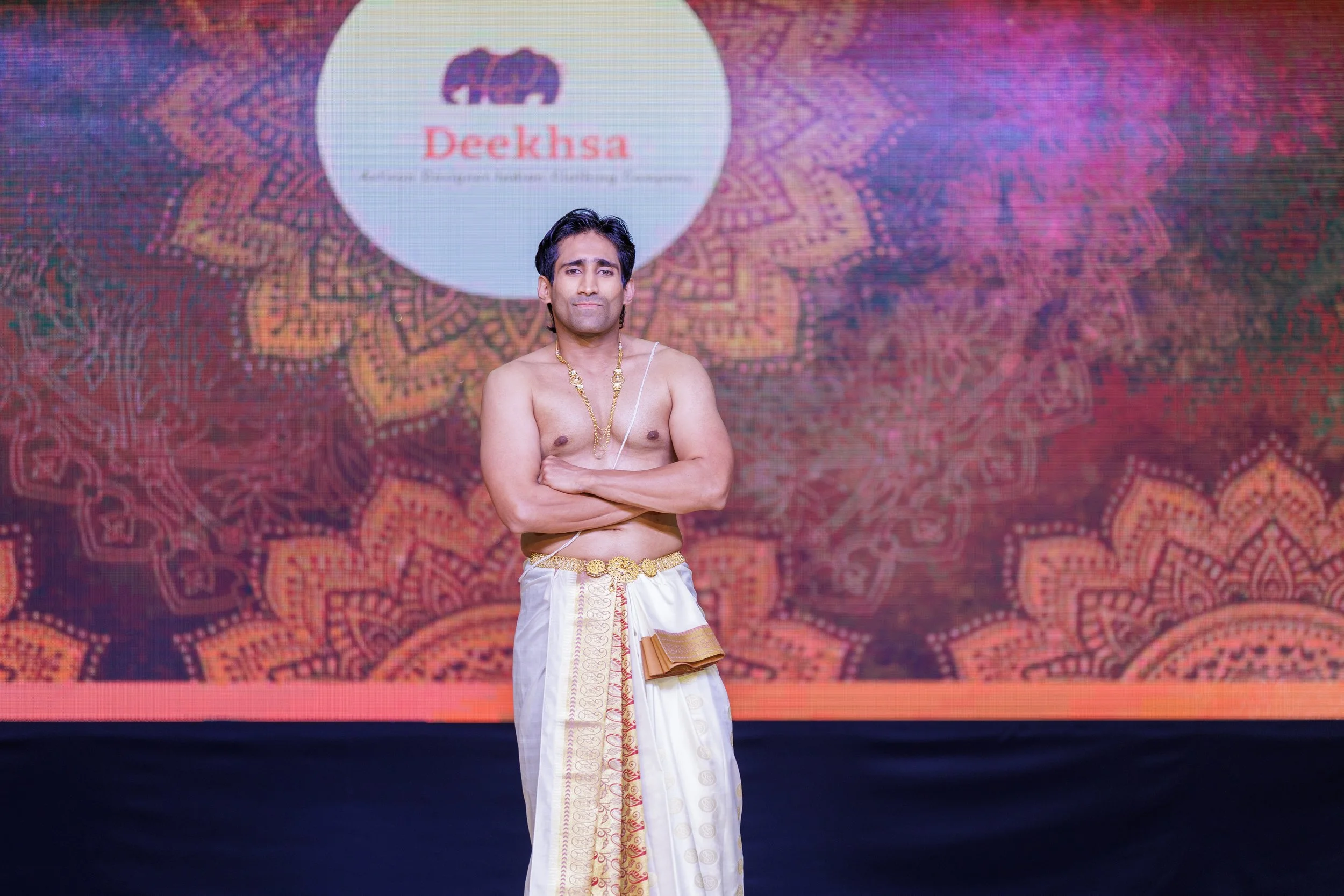A man dressed in traditional Indian attire standing on stage with a colorful intricate backdrop and a logo that reads 'Deeksha' behind him.