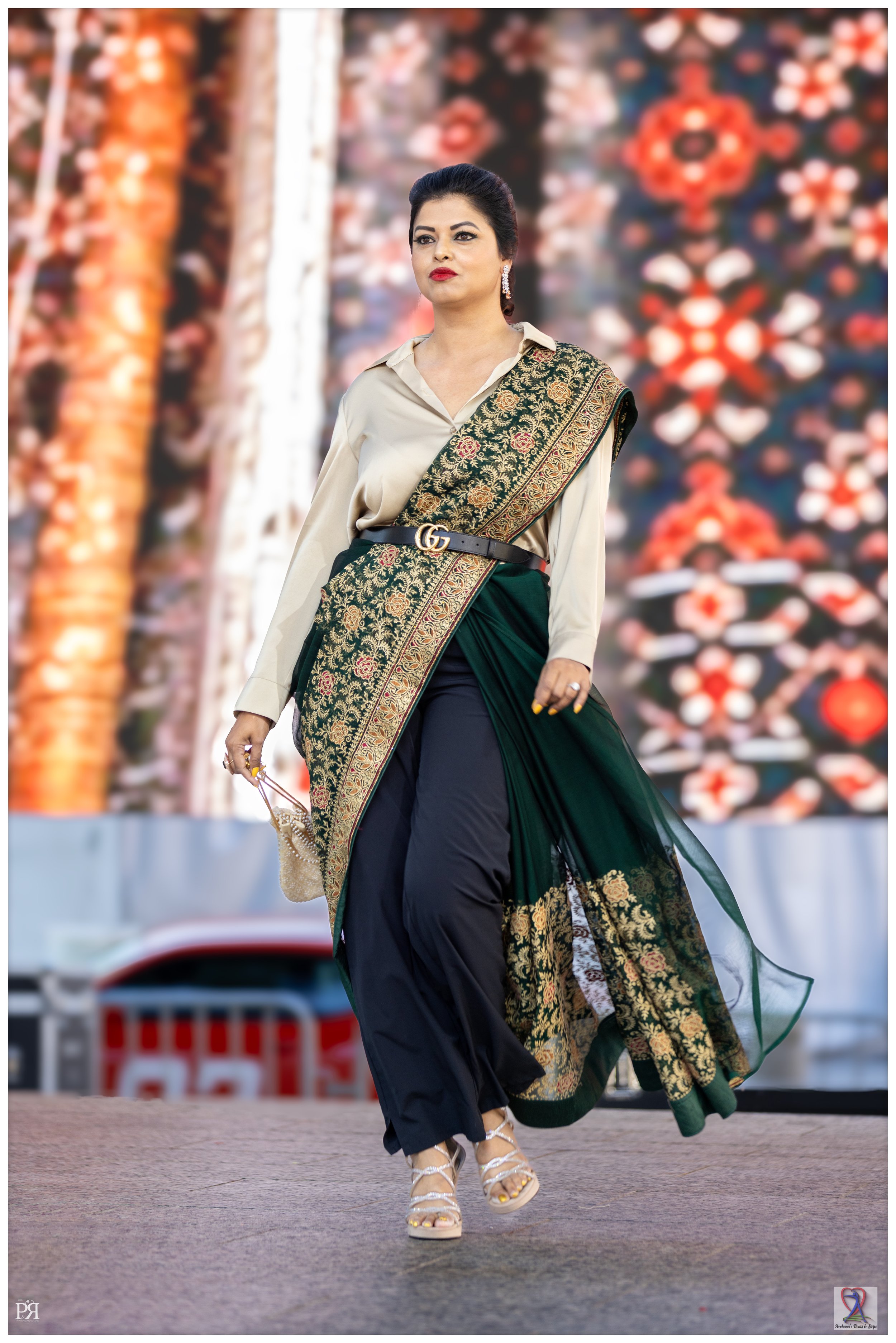 A woman is walking on a runway in a blend of traditional and modern fashion, featuring a designer belt, dangle earrings, and strappy sandals, with a colorful patterned backdrop.