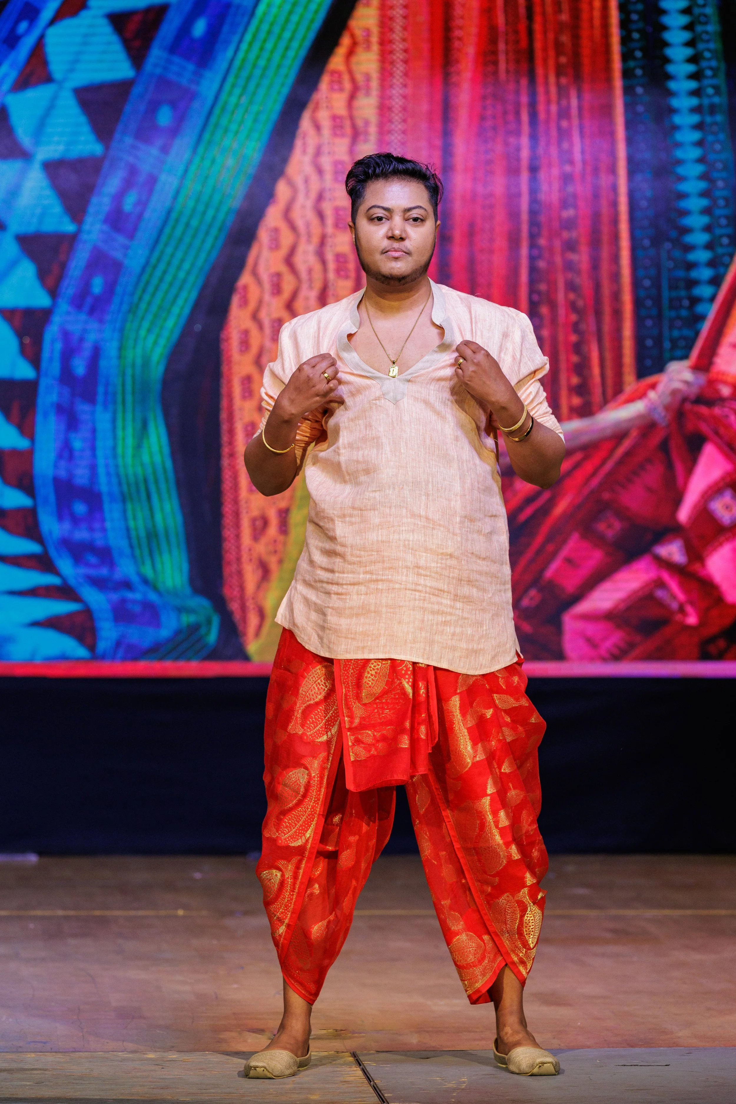 Person standing on stage in traditional attire, with colorful patterned backdrop.