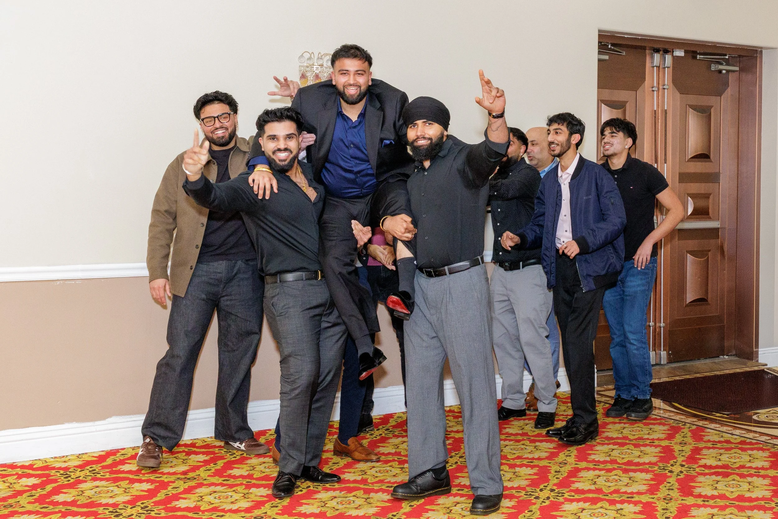 Group of men dressed in formal and casual attire celebrating indoors, some making gestures and smiling.