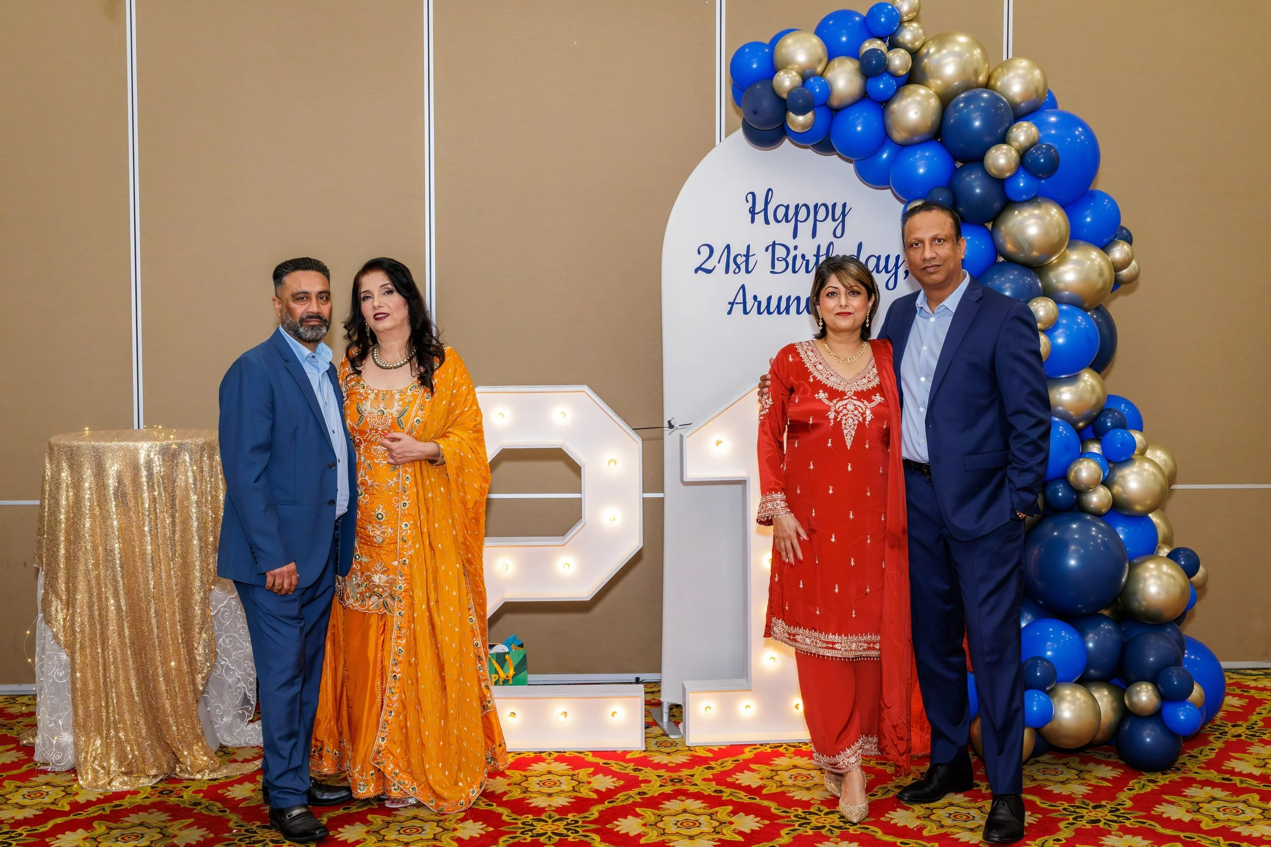 Group of four people at a birthday celebration, two men and two women dressed in colorful traditional attire, standing in front of a sign decorated with balloons that reads 'Happy 21st Birthday Aruna', and illuminated number '21' on the floor.