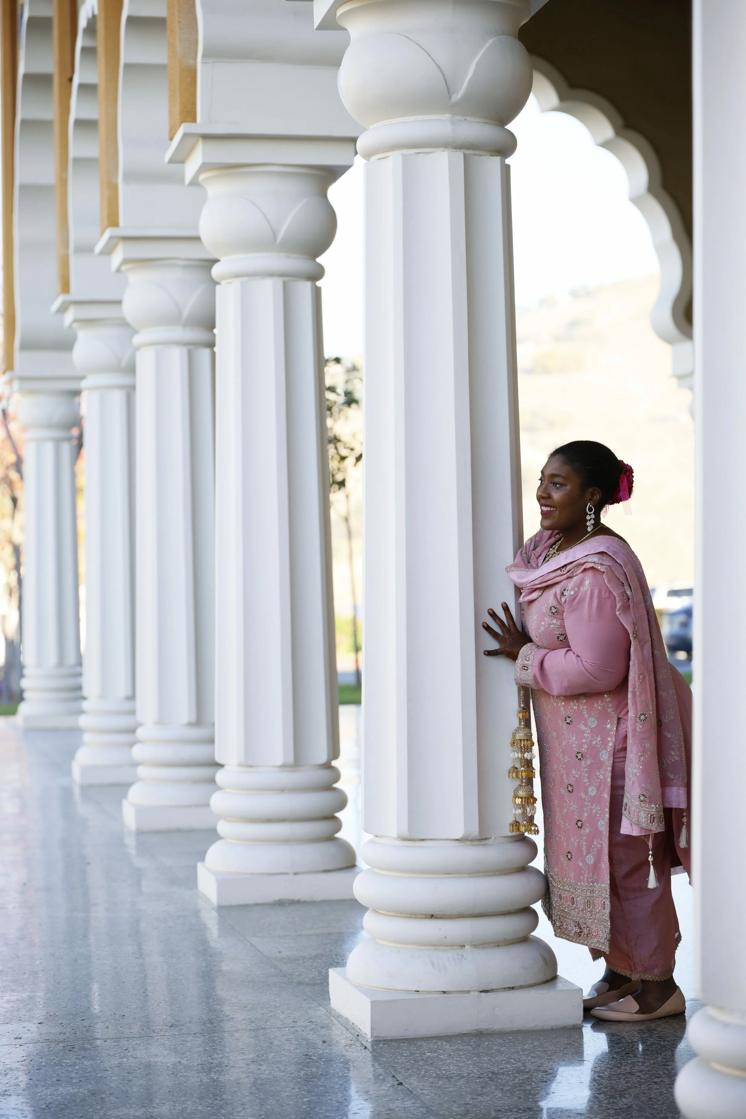 A woman dressed in traditional pink Indian attire standing behind white ornate columns on a porch, smiling and looking to the side.