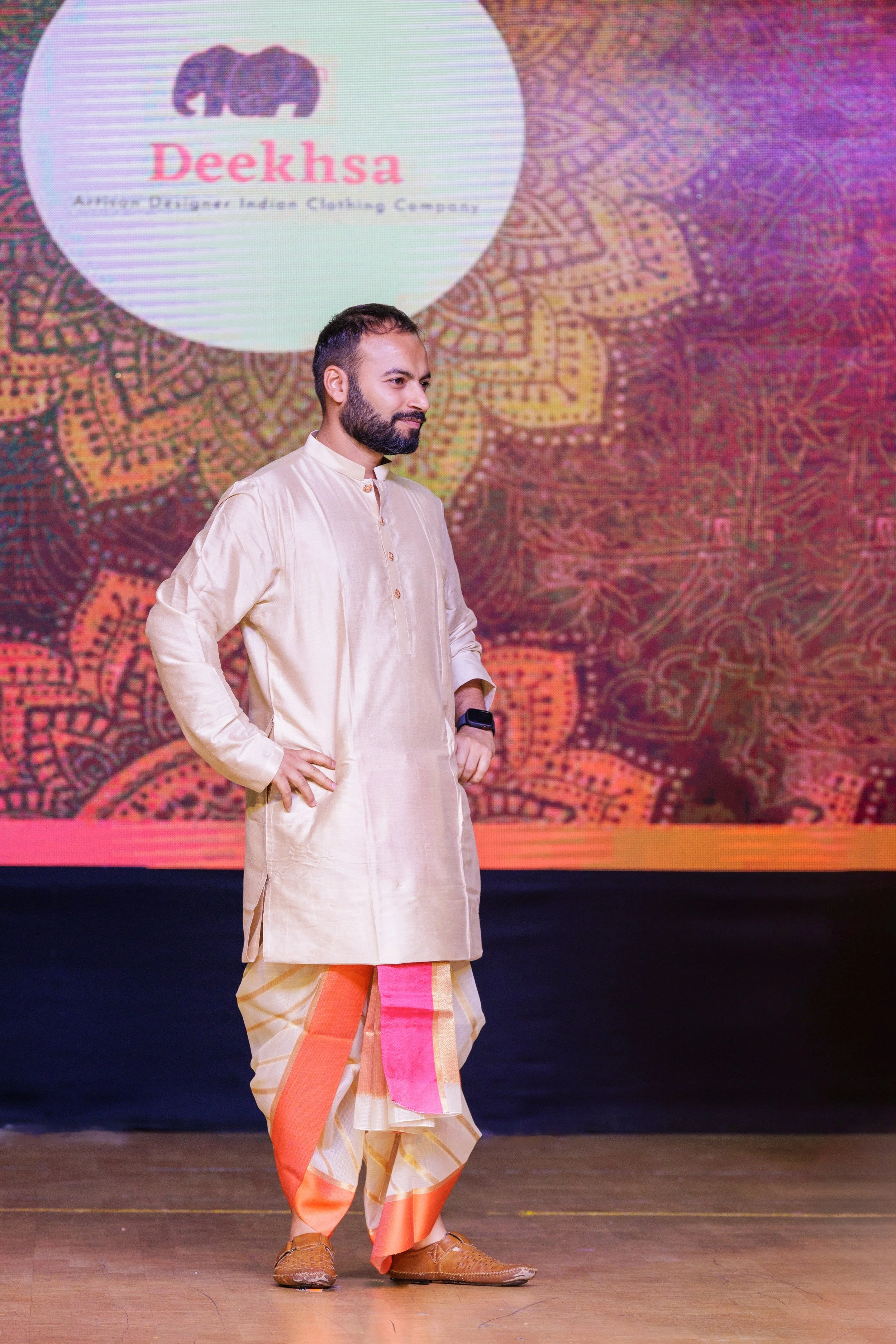 A man in traditional Indian attire, including a cream kurta and patterned dhoti, is standing on a stage with one hand on his hip. Behind him is a large screen displaying a logo for Deeksha, with a colorful, intricate background design.