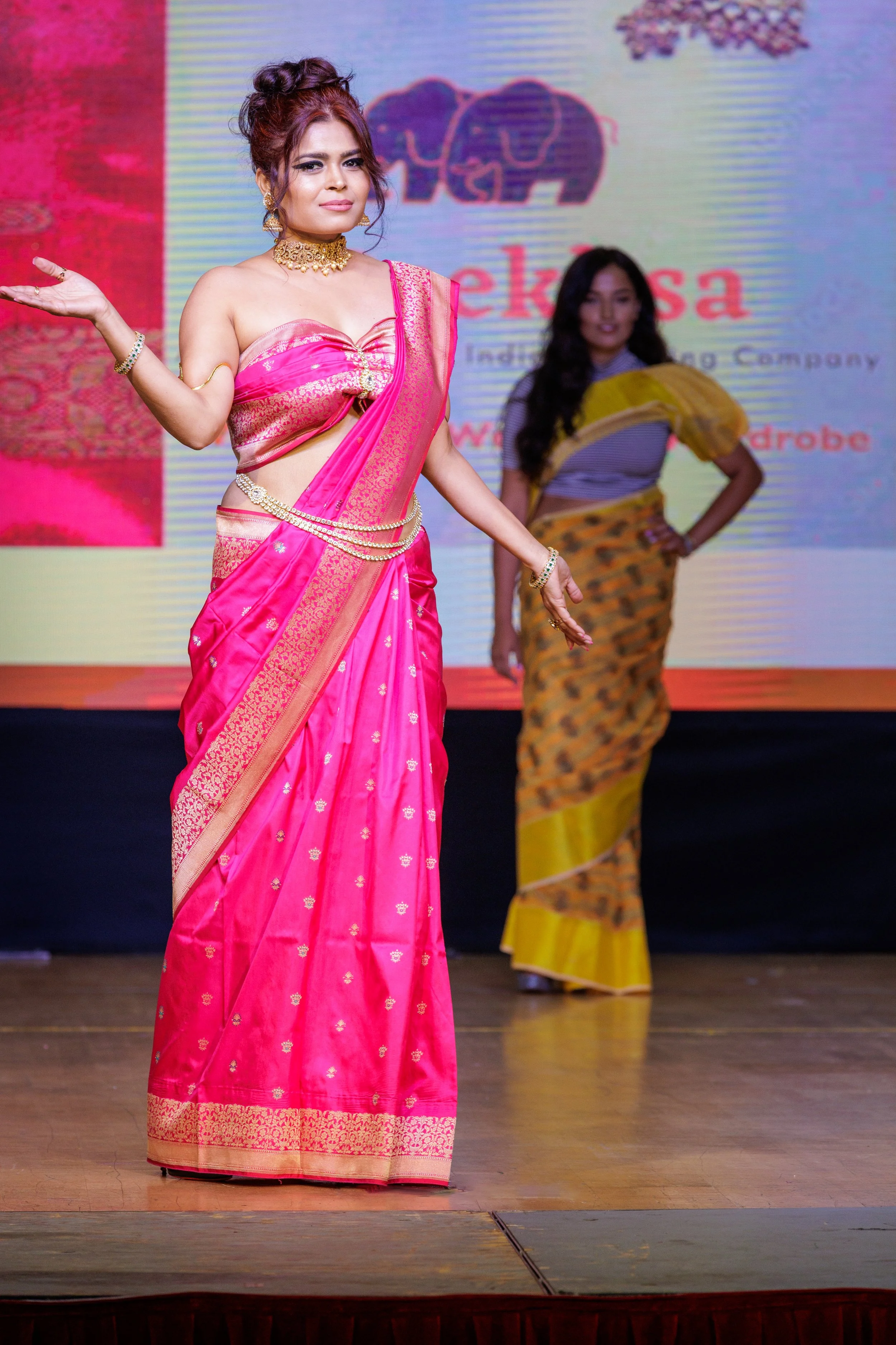 Two women wearing traditional Indian sarees on stage during a fashion show or cultural event, with a backdrop displaying a logo and text.