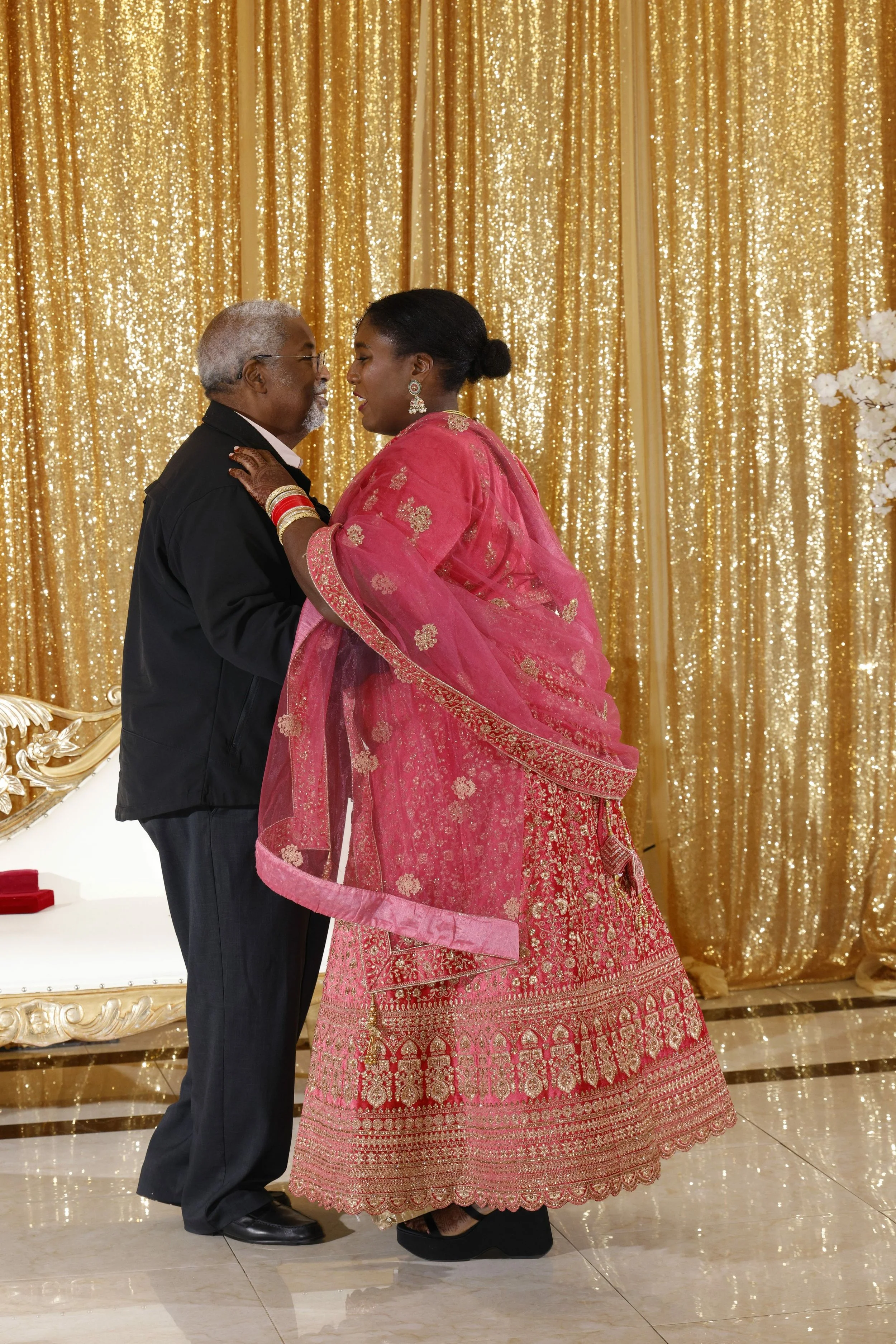 An elderly man and woman dancing together at a celebration, with a golden, glittery backdrop and white floral decorations.
