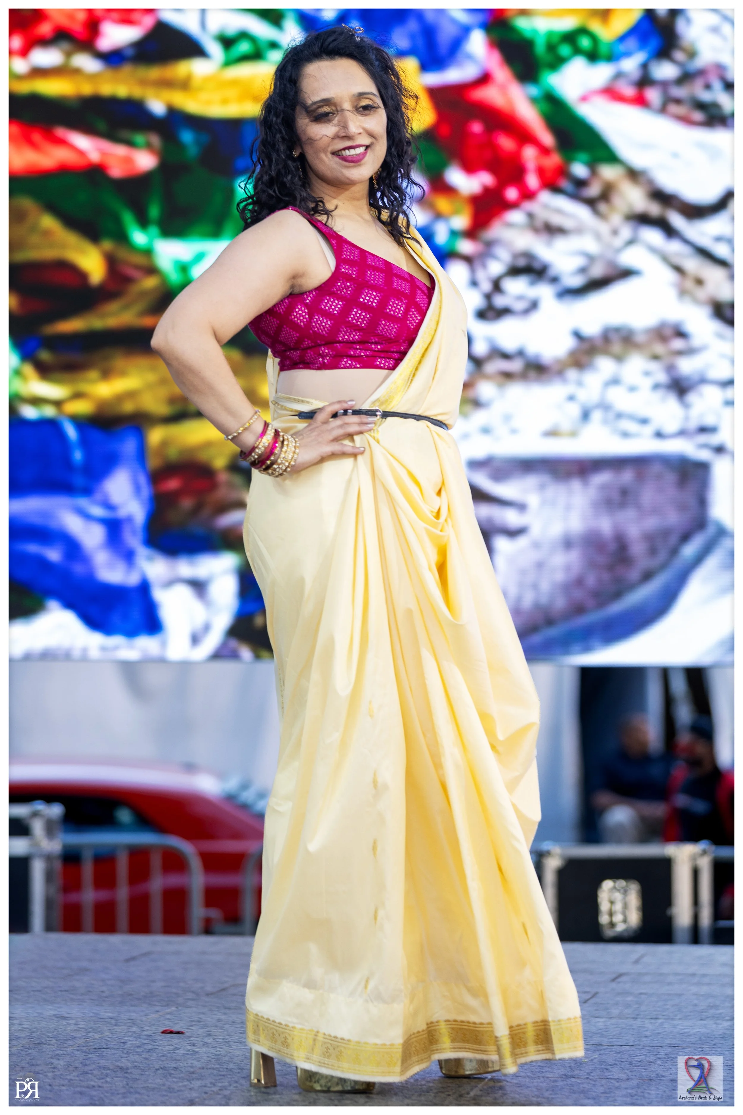 A woman is standing on stage wearing a pink blouse and a yellow saree, with a colorful background behind her.