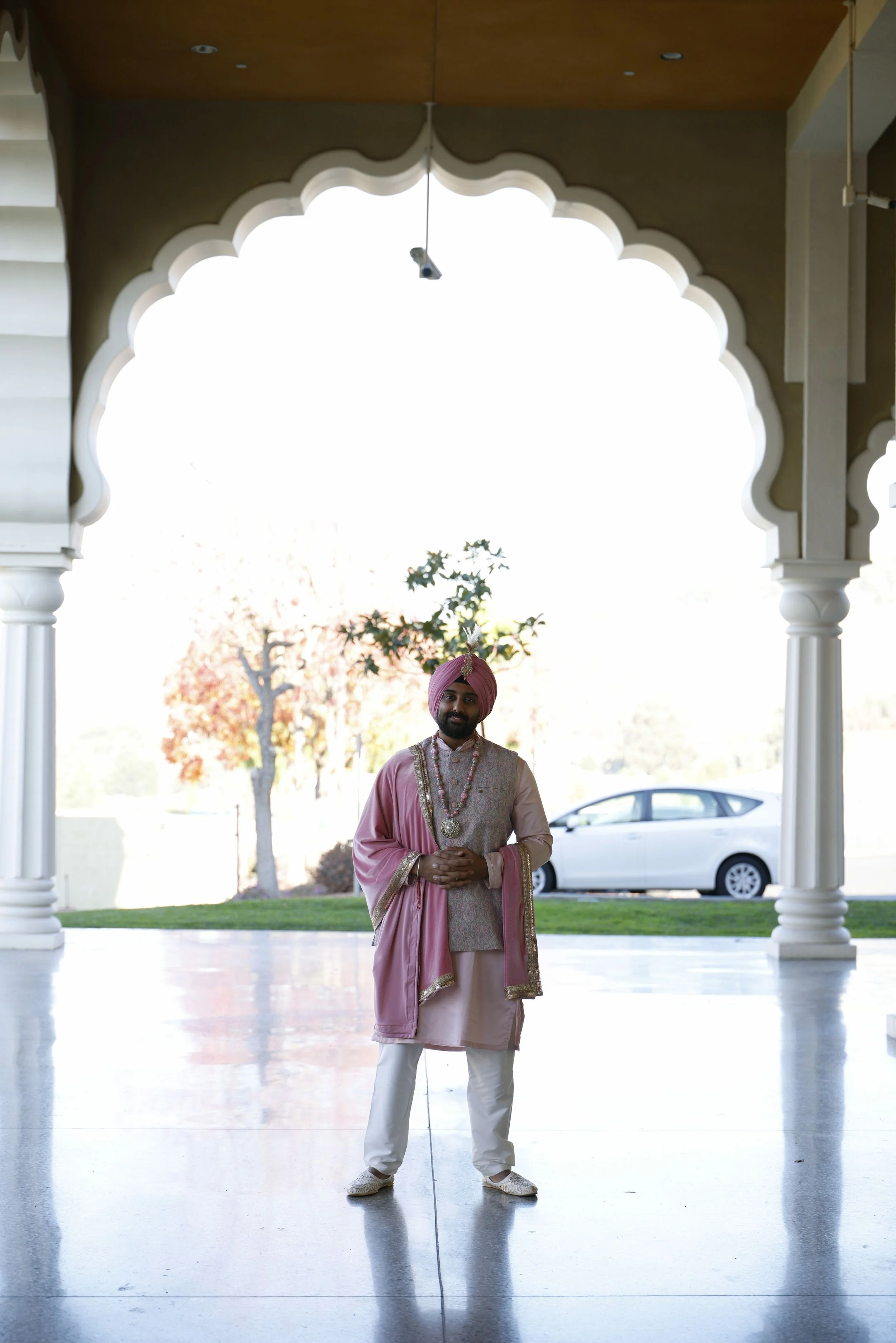 A man in traditional Indian attire, wearing a pink turban, pink and beige embroidered outfit, and jewelry, standing under an ornate archway with his hands clasped, smiling, with trees and a car in the background.
