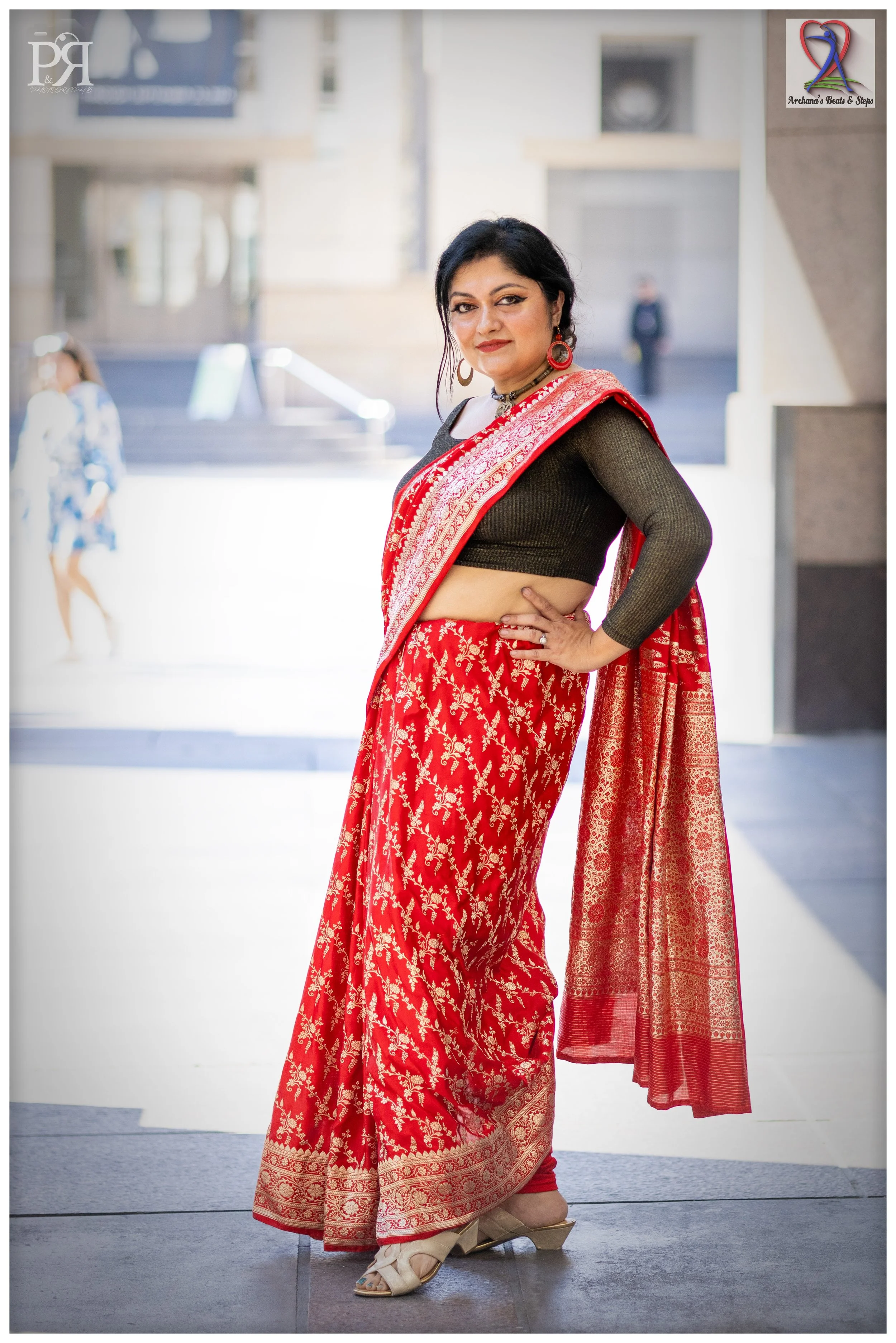 A woman in a black crop top and red saree with gold embroidery, standing outdoors with her hand on her hip, smiling at the camera.