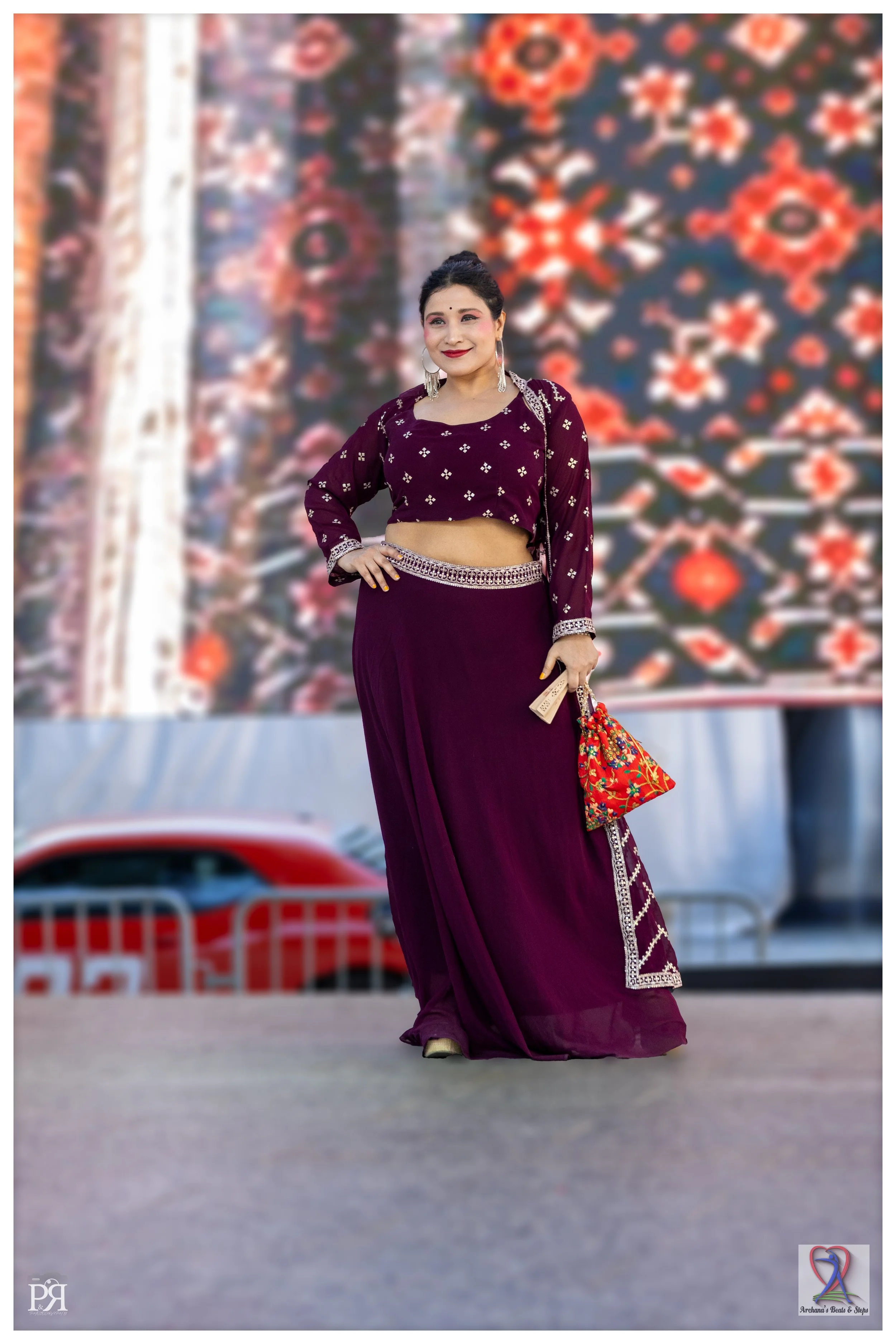 A woman wearing traditional Indian attire, a deep purple lehenga with a matching blouse, standing outdoors in front of a colorful, ornate patterned backdrop. She is smiling, holding a small, colorful handbag, and has jewelry and makeup on.