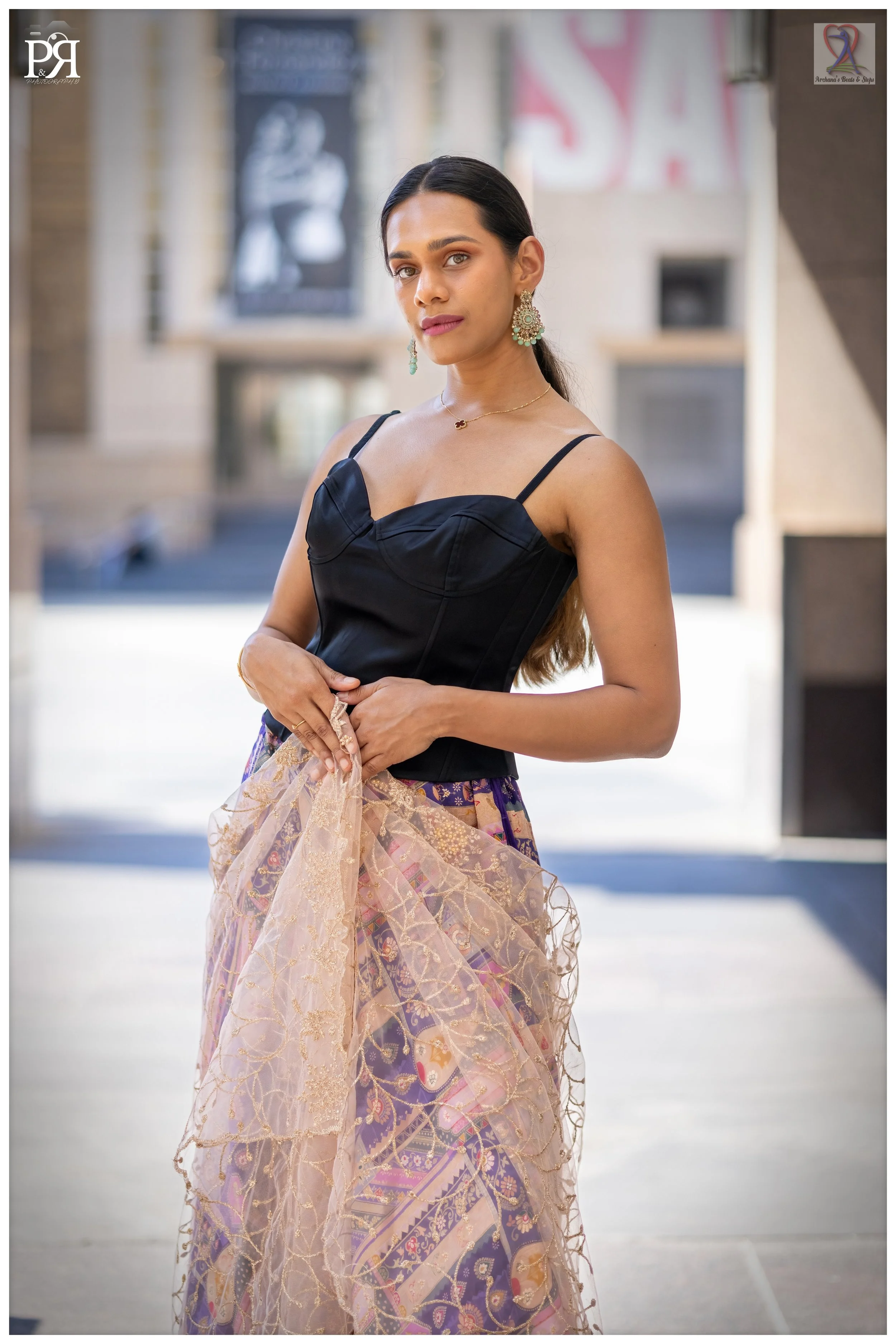 A young woman stands outdoors in a city setting wearing a black sleeveless top and a colorful skirt with intricate patterns, holding a sheer, gold-embroidered fabric. She has long dark hair, earrings, and a delicate necklace.