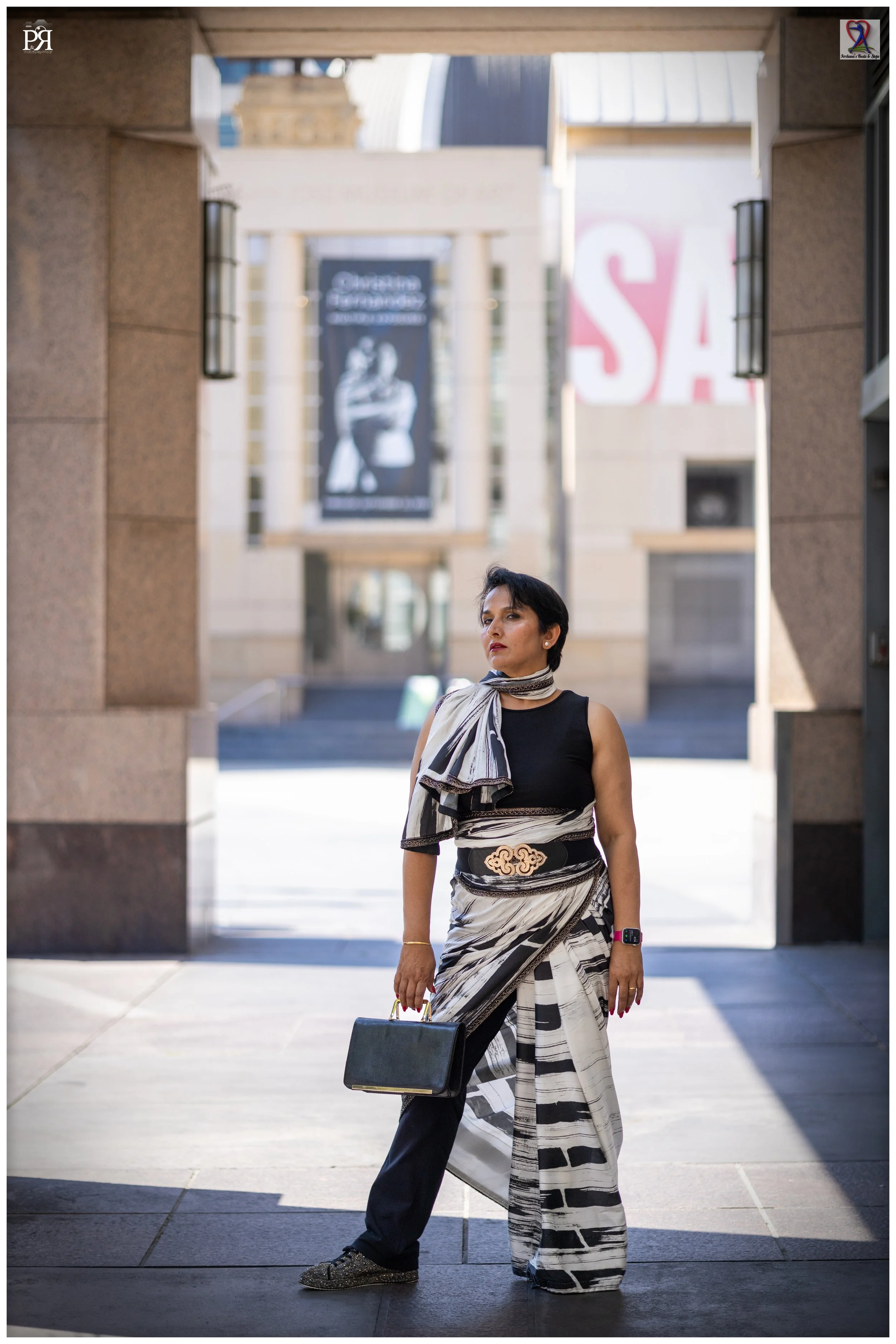 A woman standing outdoors in front of a building with large banners, wearing a black and white patterned ensemble, holding a black handbag, and looking to the side.