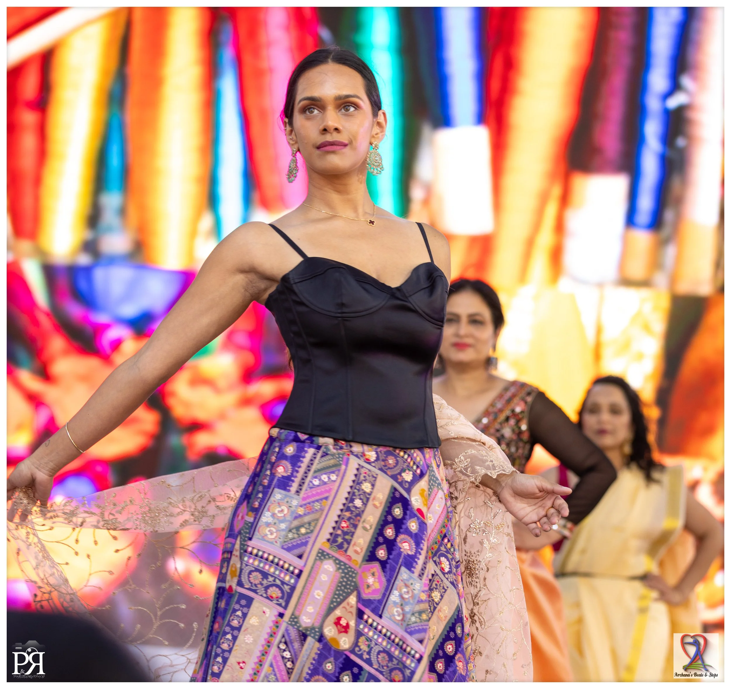 Woman in a black corset top and vibrant, patterned skirt, dancing at an indoor event with a colorful backdrop.