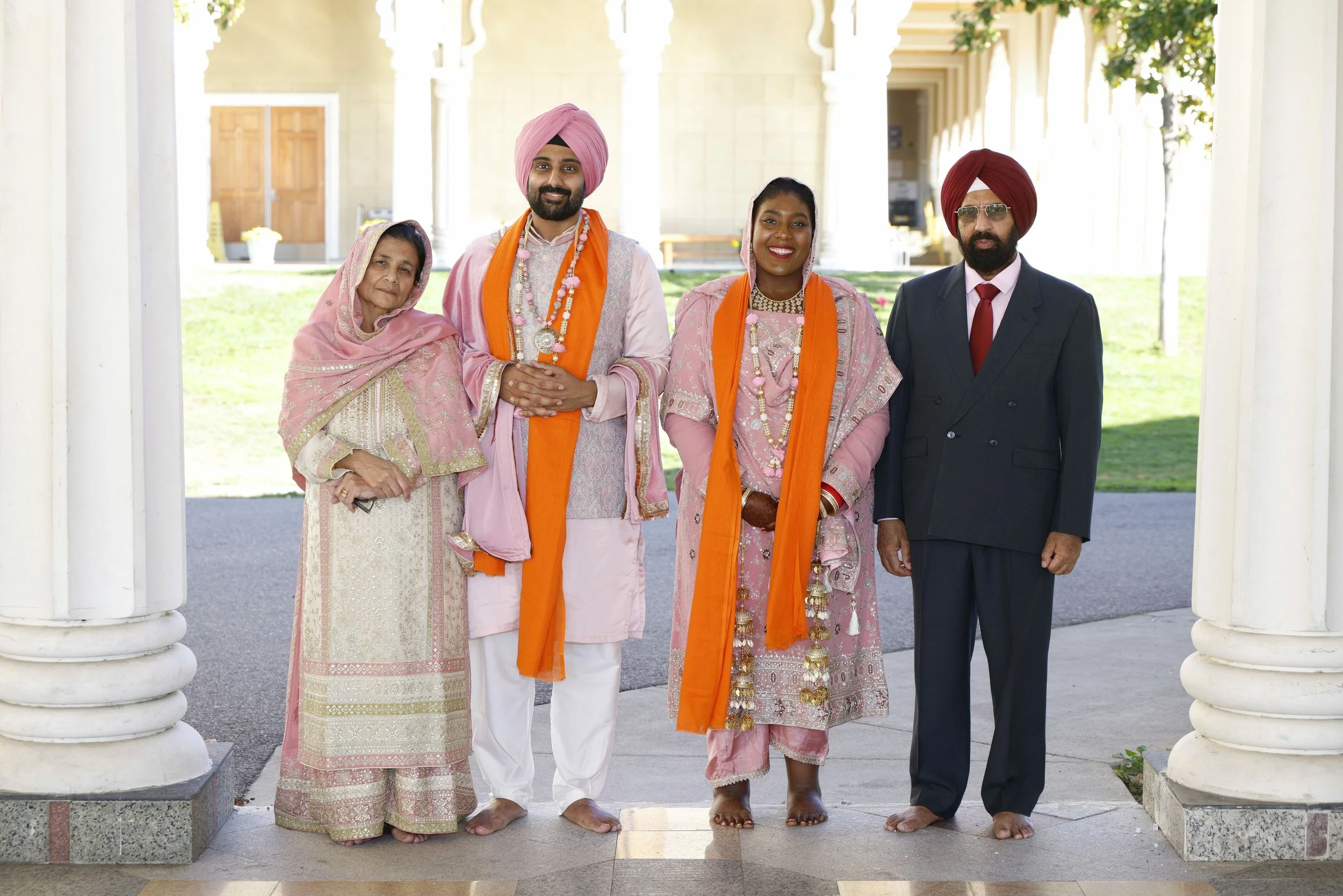 Five people standing outdoors under a white archway, likely at a wedding or cultural celebration, dressed in traditional Indian attire with bright colors, jewelry, and turbans.