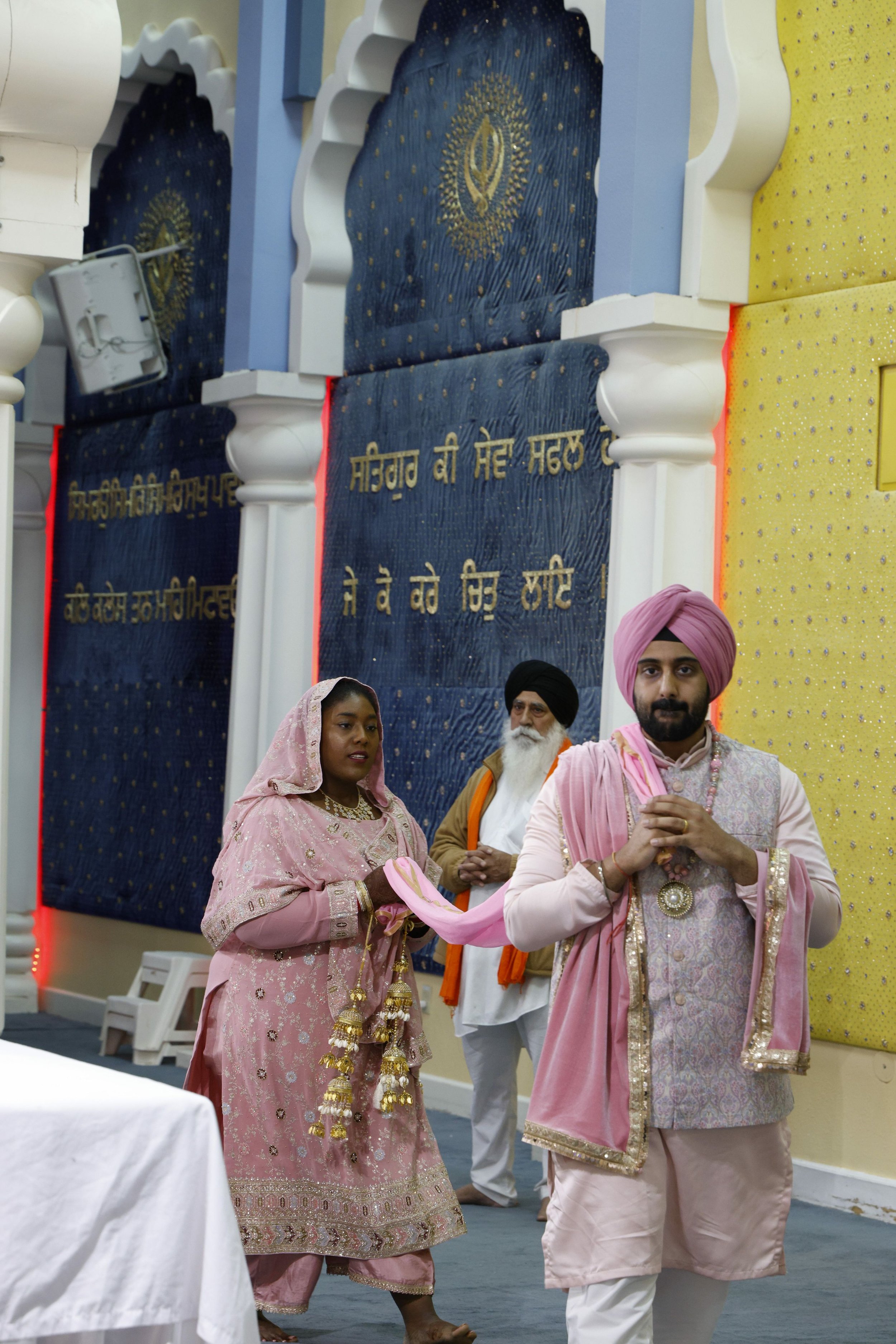 A Sikh wedding ceremony with the bride and groom dressed in traditional pink and cream attire, standing in front of a decorated Gurdwara interior with blue and yellow wall hangings and gold-scripted Punjabi text, accompanied by an older man in the ba