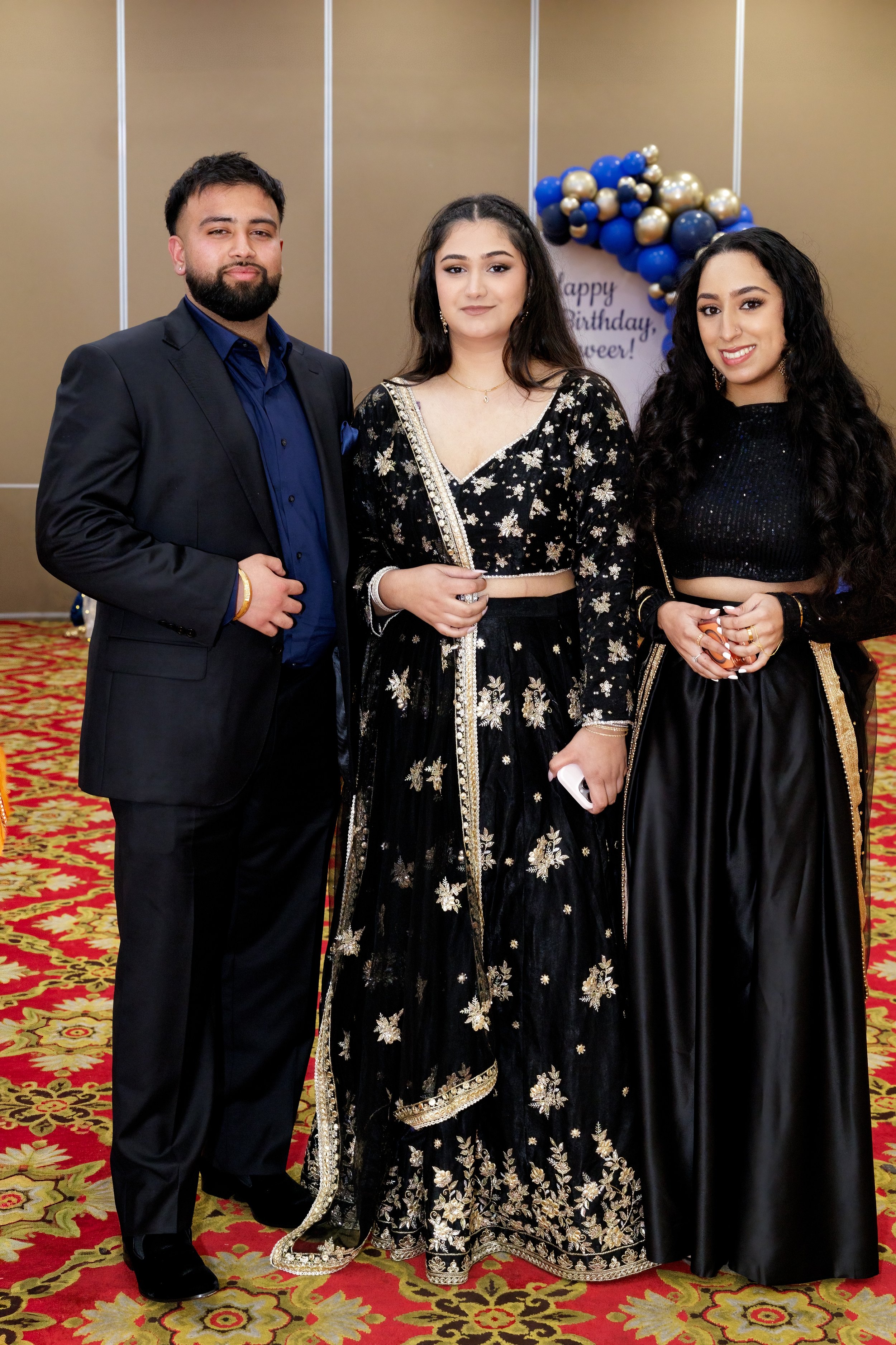 Three young adults dressed in formal attire at a celebration, standing on a red and gold patterned carpet, with a decorated backdrop featuring a wonky balloon arrangement and a sign that reads "Happy Birthday," in an indoor event space.