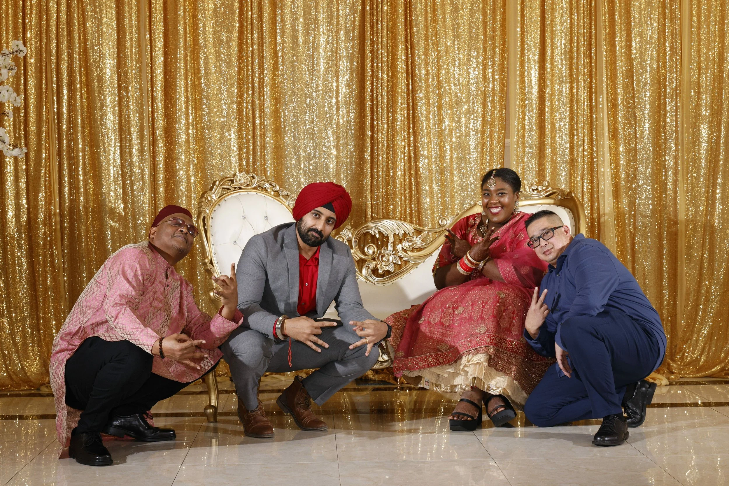 Four people at a celebratory event, with a gold sequin backdrop and an ornate white and gold throne, two men kneeling and two women sitting, dressed in traditional and modern attire, smiling and making gestures.