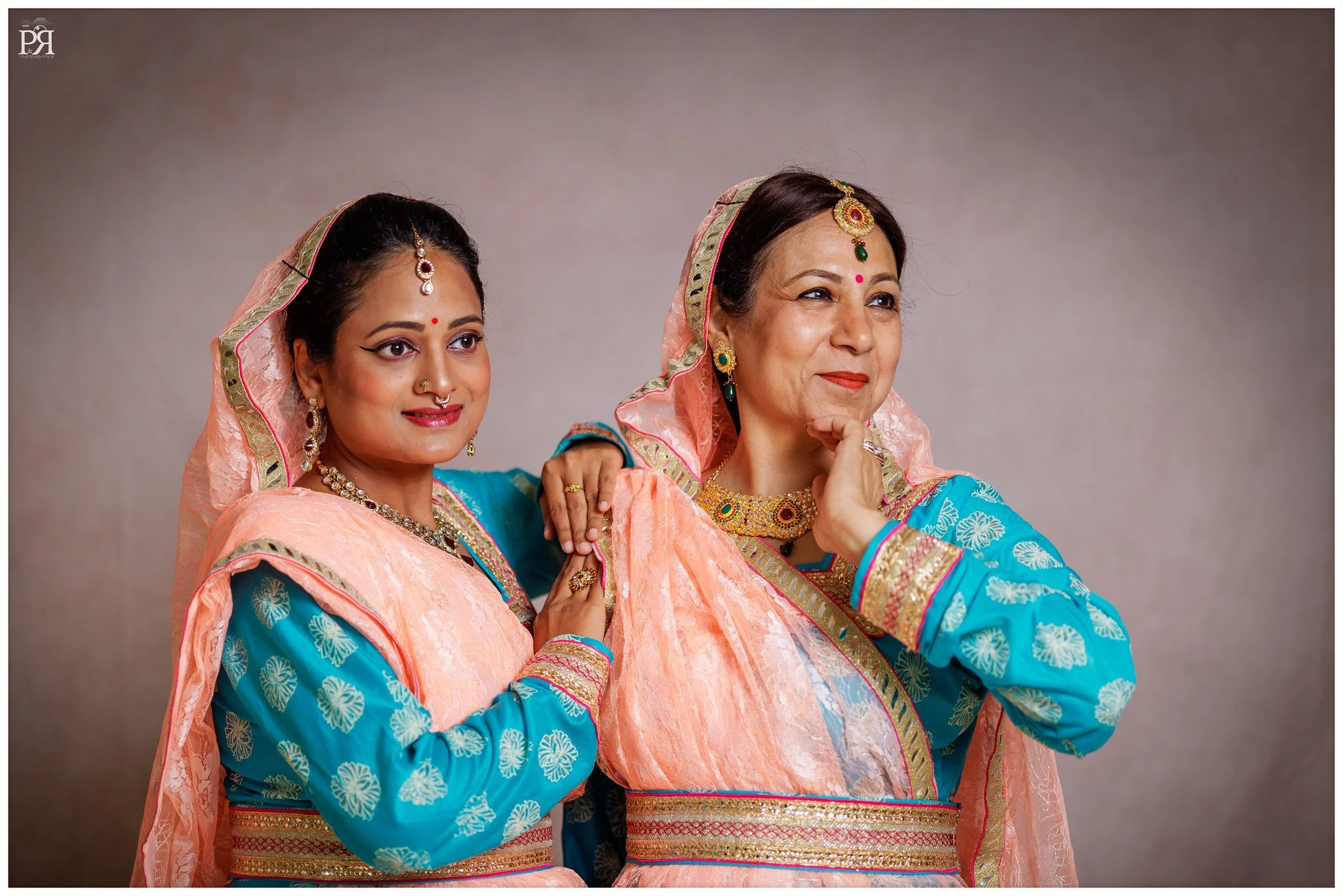 Two women dressed in traditional Indian attire, smiling and posing for the camera with a neutral background.