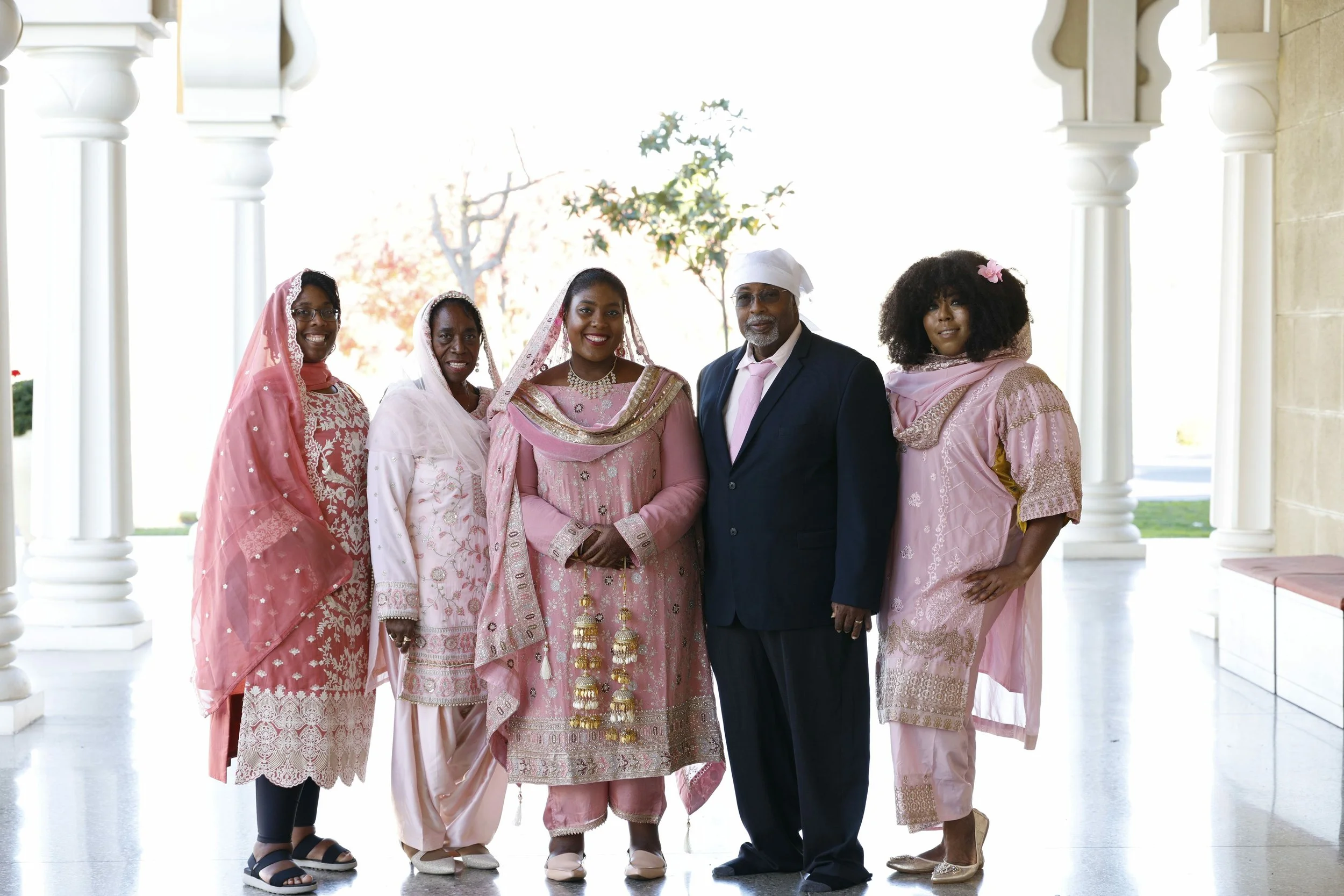Group of six people standing together in traditional and formal attire, smiling on a bright porch with columns.