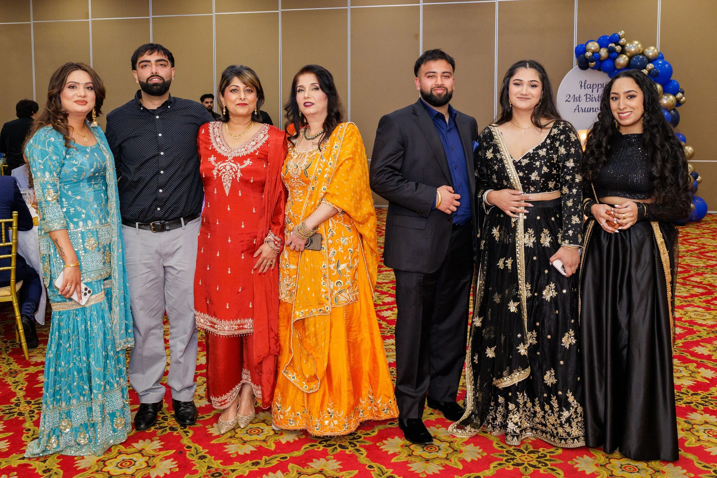 Group of people dressed in traditional Indian attire, standing on a patterned carpet in a decorated event space with a blue and gold balloon arch in the background.
