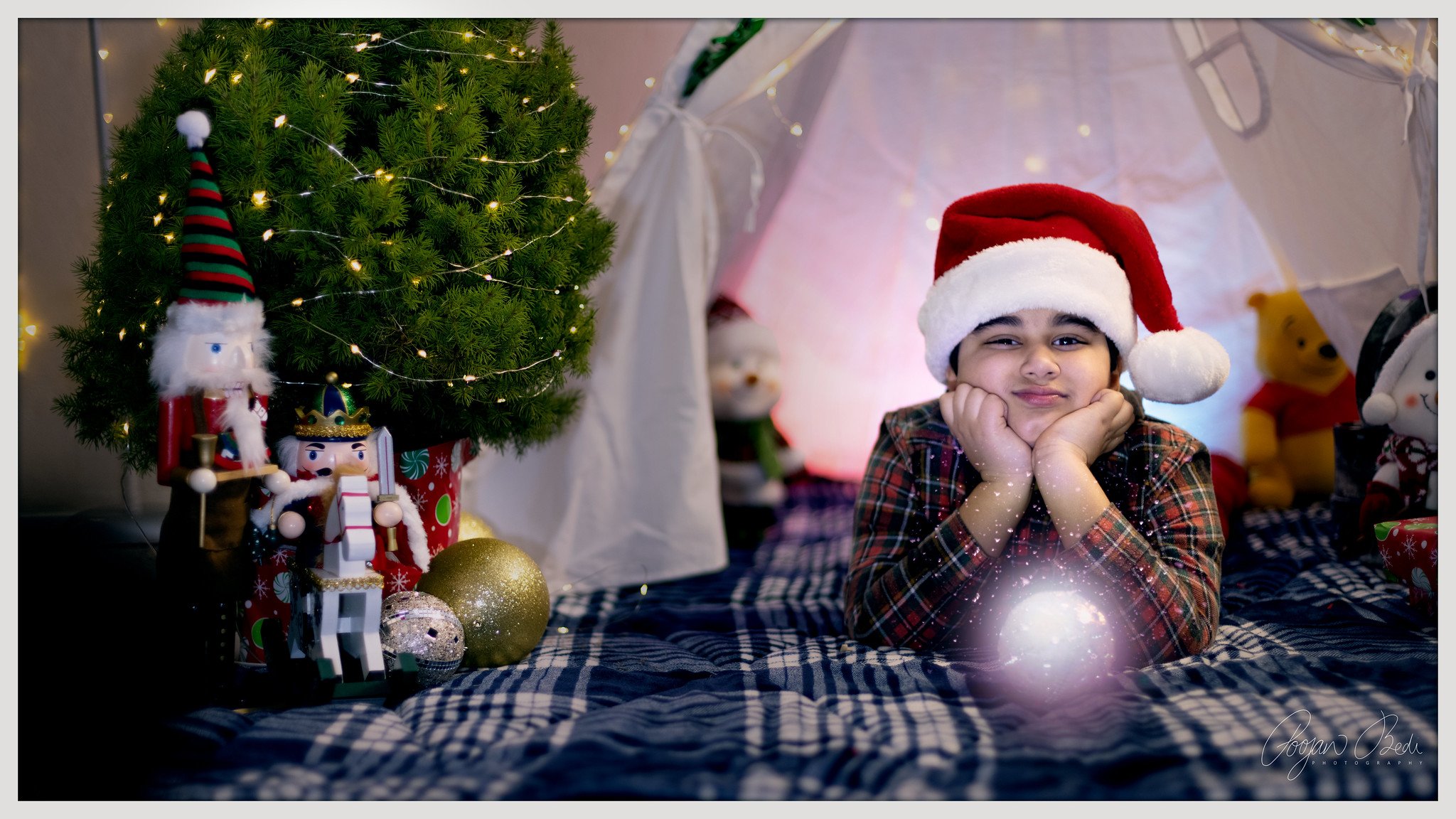 A young boy lying on a bed with a checkered blanket, wearing a Santa hat, resting his chin on his hands, surrounded by Christmas decorations including a decorated Christmas tree with lights, ornaments, and stuffed toys like Winnie the Pooh and Cloud.