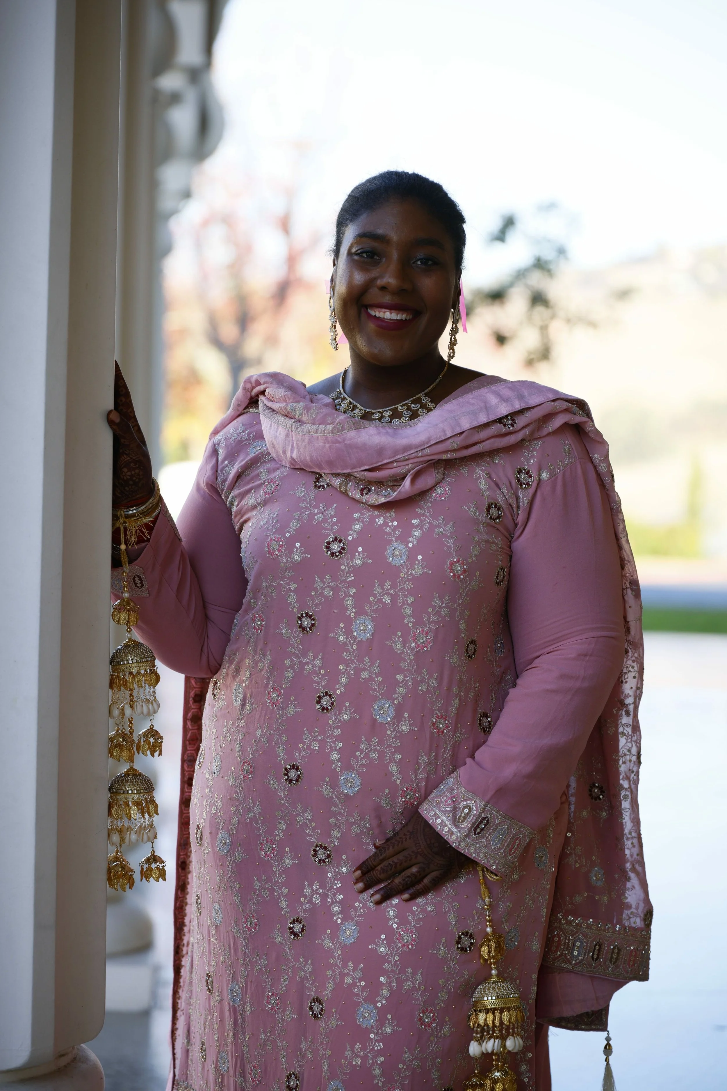 A woman dressed in traditional pink embroidered attire with jewelry, smiling outdoors, standing near columns with trees and sky in the background.