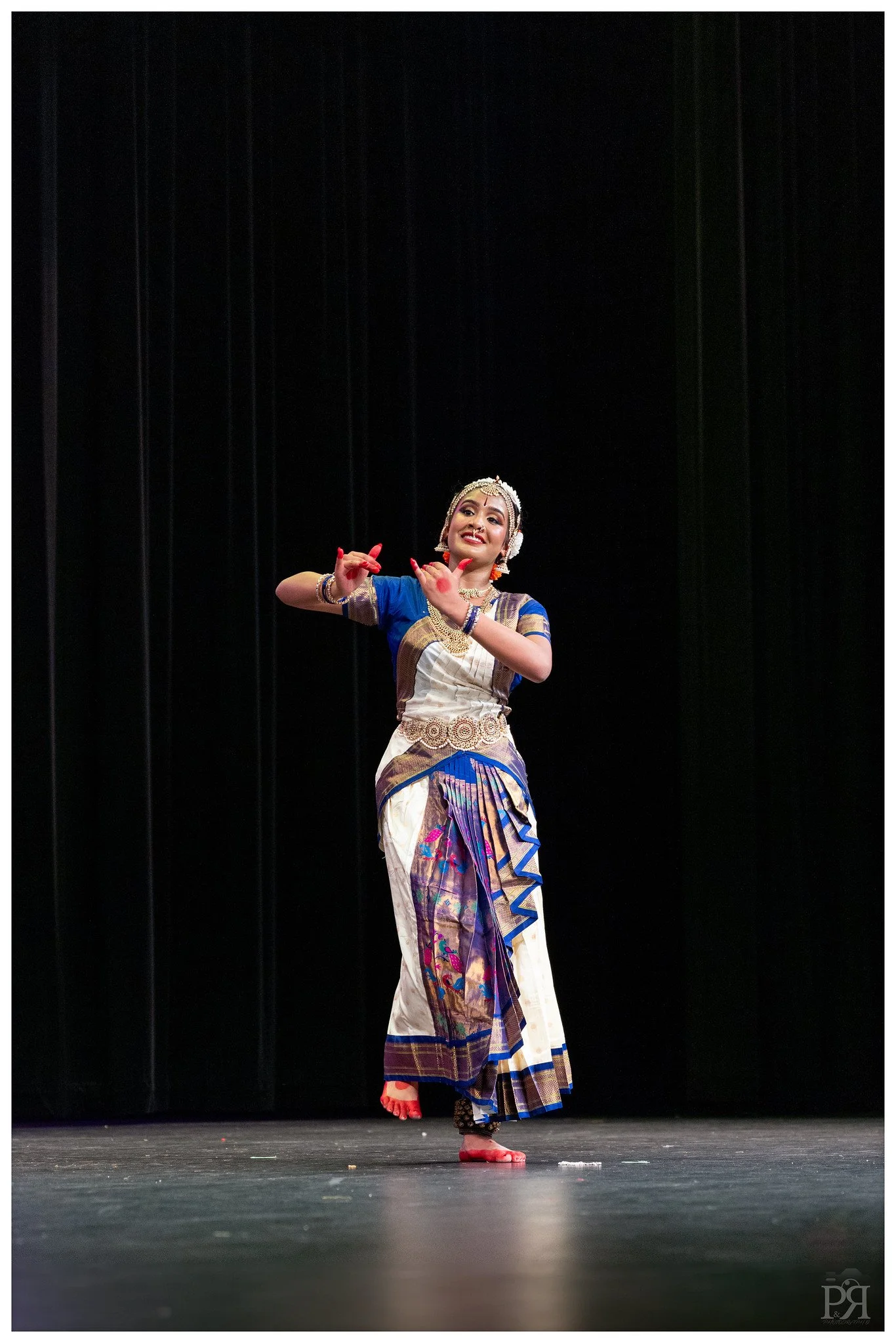 A woman in traditional Indian dance attire performing on stage, wearing a white and blue sari with gold accents, jewelry, and makeup, against a black background.