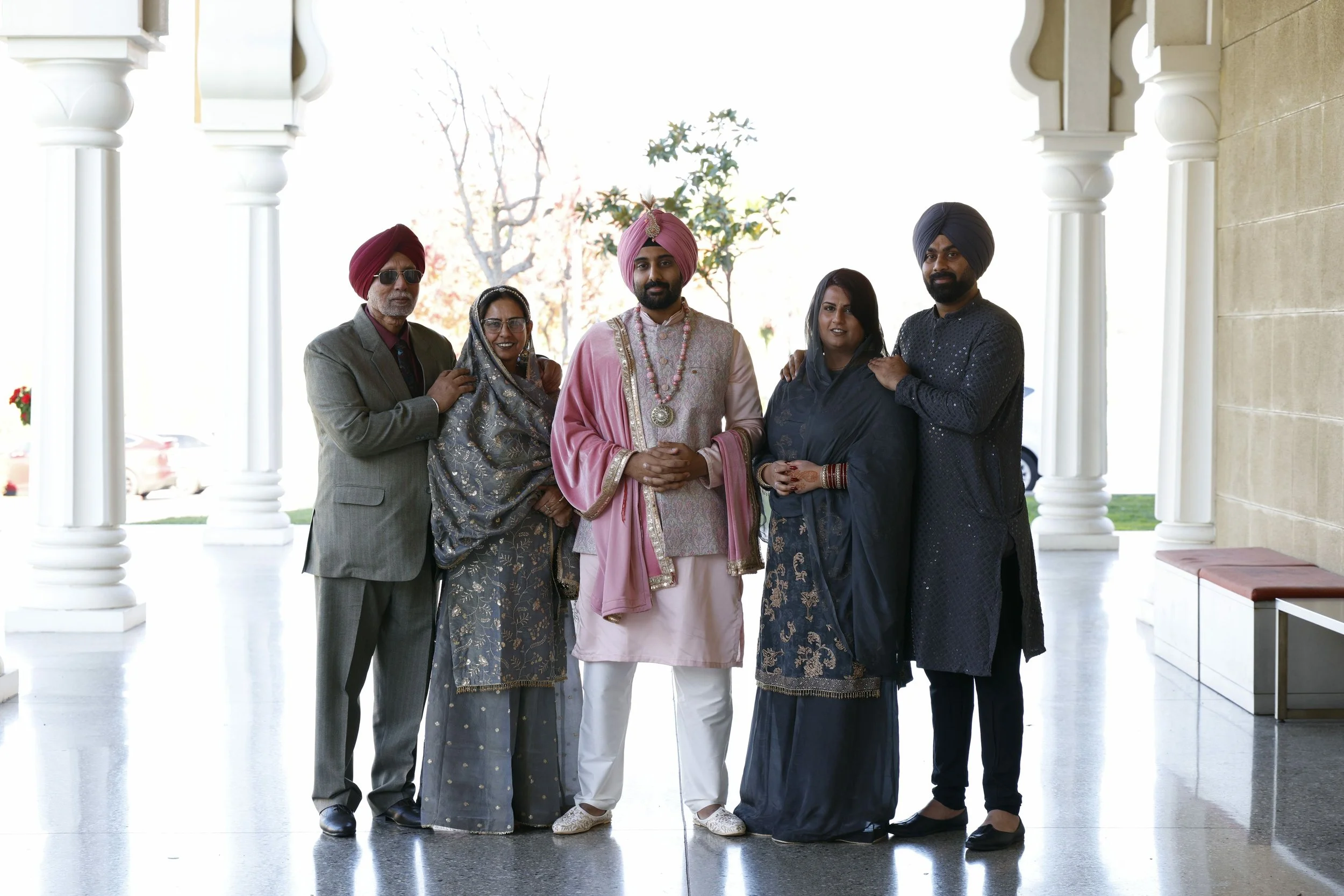 Family portrait of six people with four men and two women, in traditional Indian attire, standing under a decorative arch, with trees in the background.