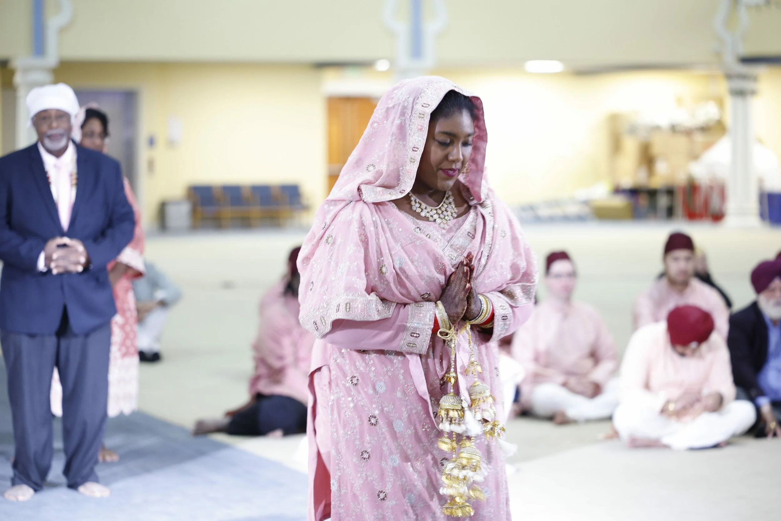 A woman in traditional Indian attire, pink saree, with jewelry and henna on her hands, praying with hands folded in a temple or prayer hall, surrounded by other seated worshippers.