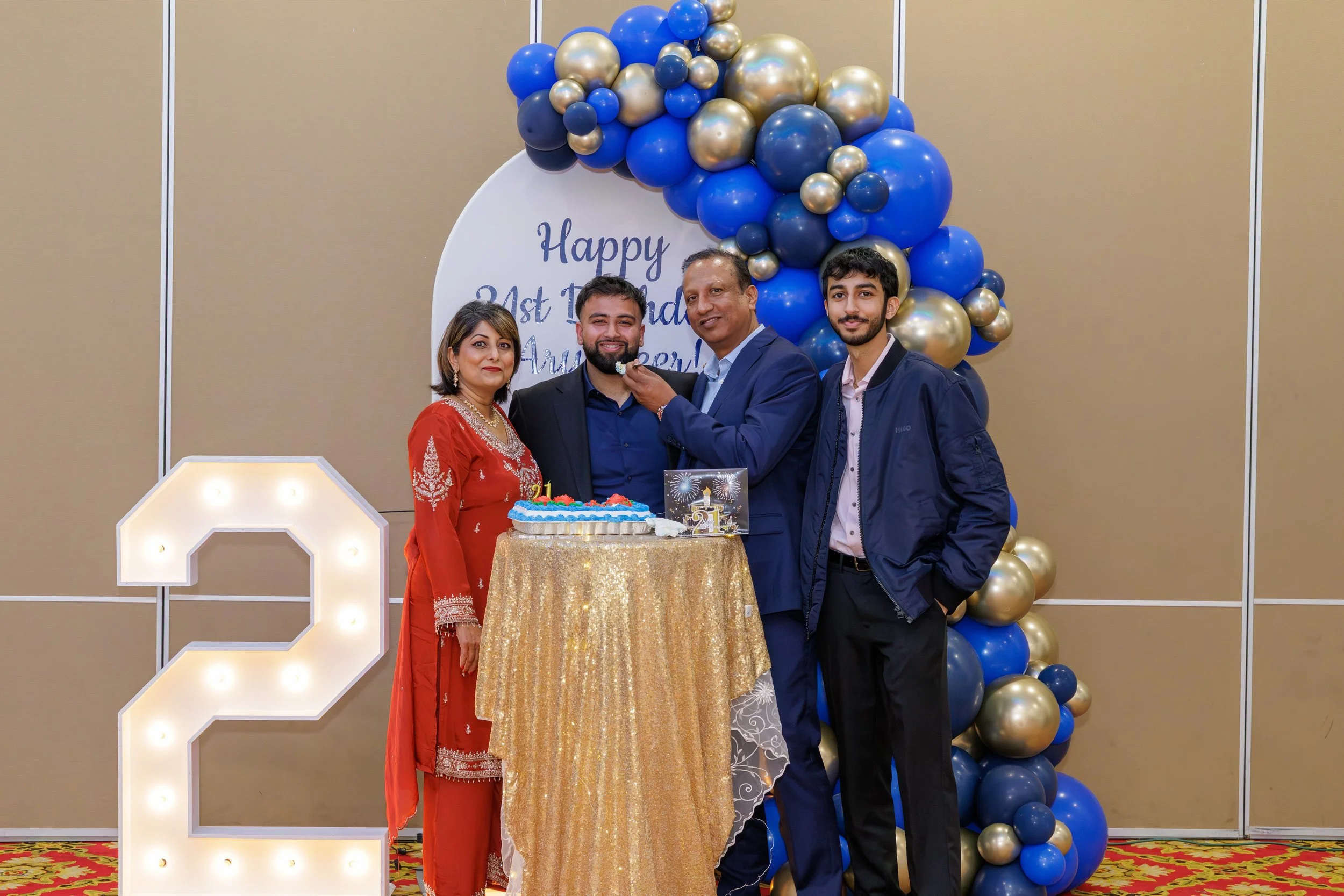 A family celebrating a 21st birthday with a decorated party scene, including a cake, a large illuminated number 2, and a balloon arch in blue, gold, and black colors, in front of a wall with a speech bubble-shaped sign that reads 'Happy 21st Birthday