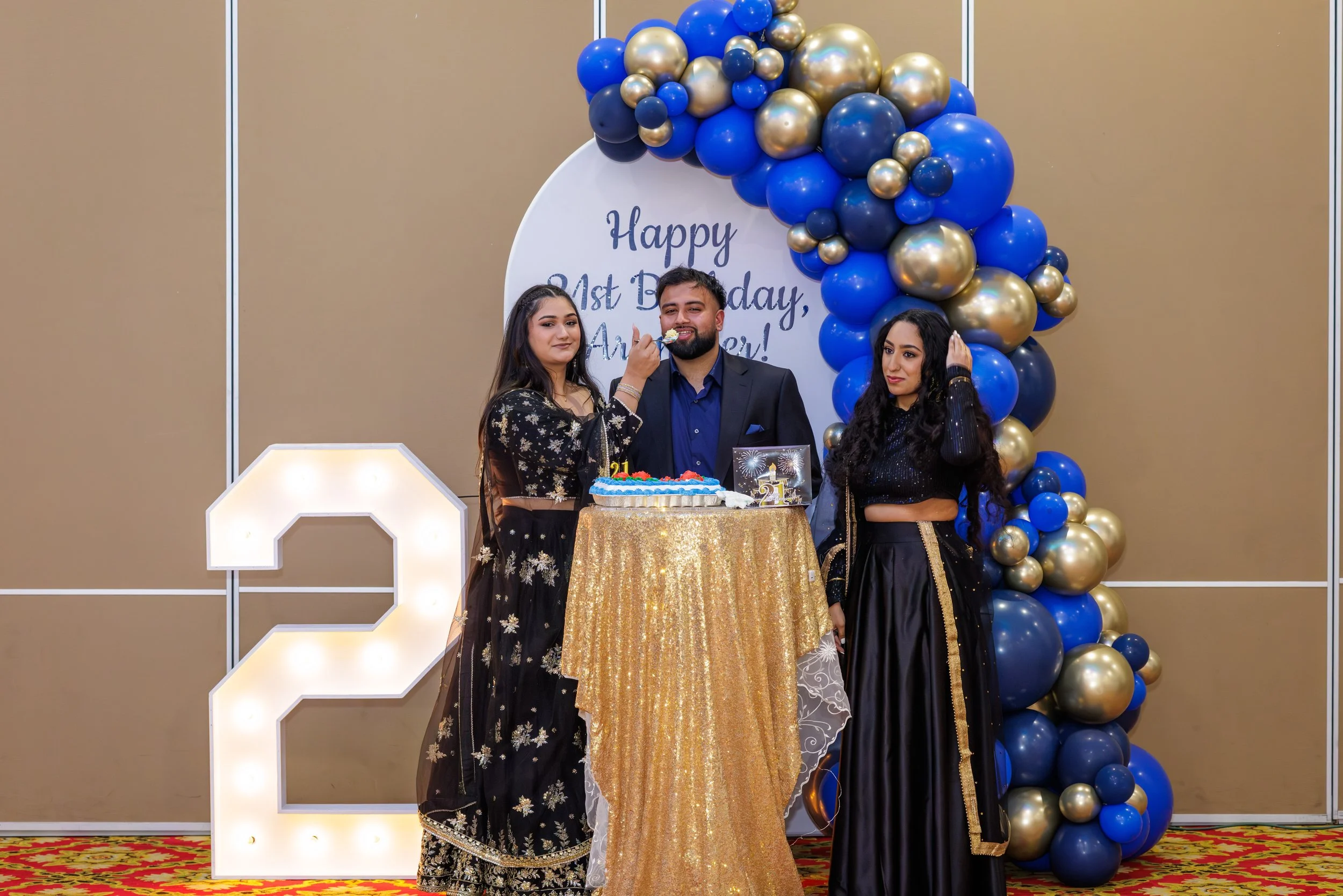 Celebrating a 21st birthday with three young adults standing around a table with a birthday cake, gold and blue balloons, a large illuminated number 21, and a backdrop that says 'Happy 21st Birthday, Amber!'