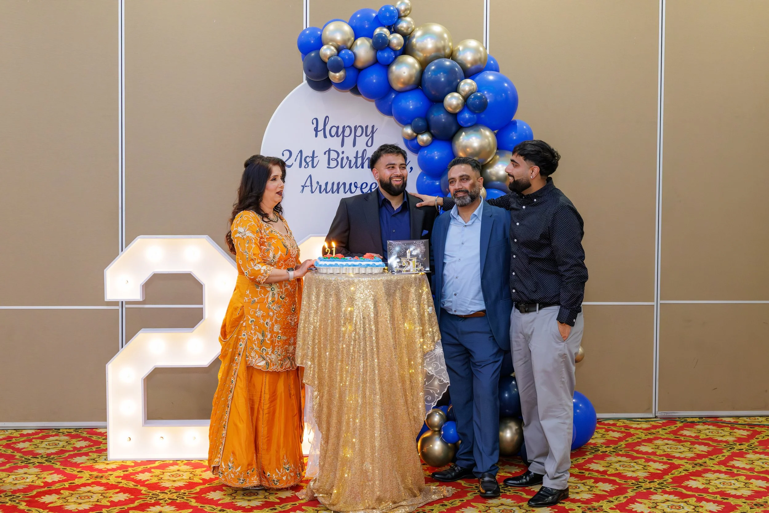 A family celebrating a 21st birthday at a party, standing around a table with a birthday cake. The background has balloons in blue and gold, and a large illuminated number 2. A woman in an orange traditional outfit holds a cake, while three men in su