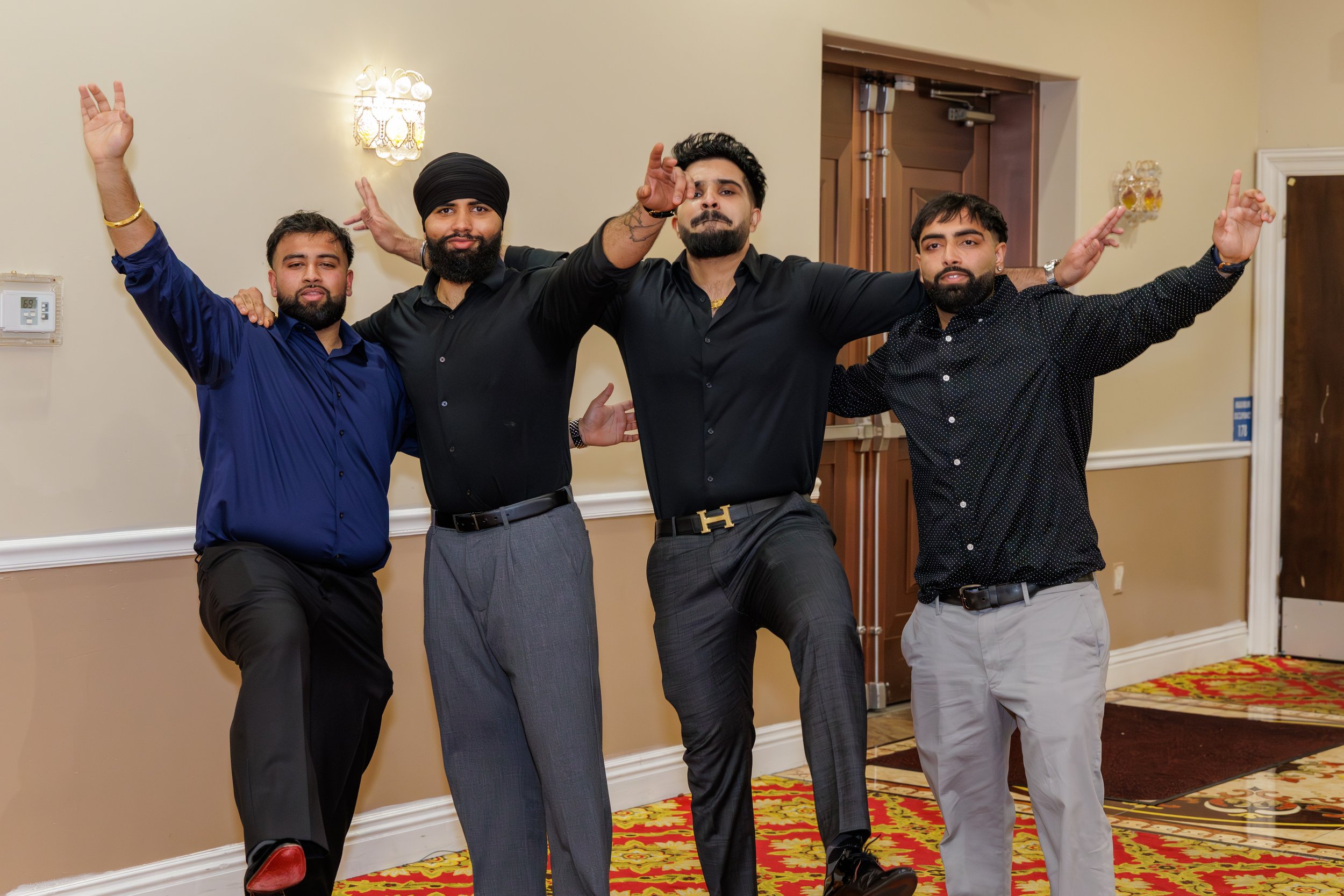 Four young men standing together indoors, celebrating, with arms outstretched or around each other, dressed in formal and semi-formal attire, with a patterned carpet and beige walls in the background.