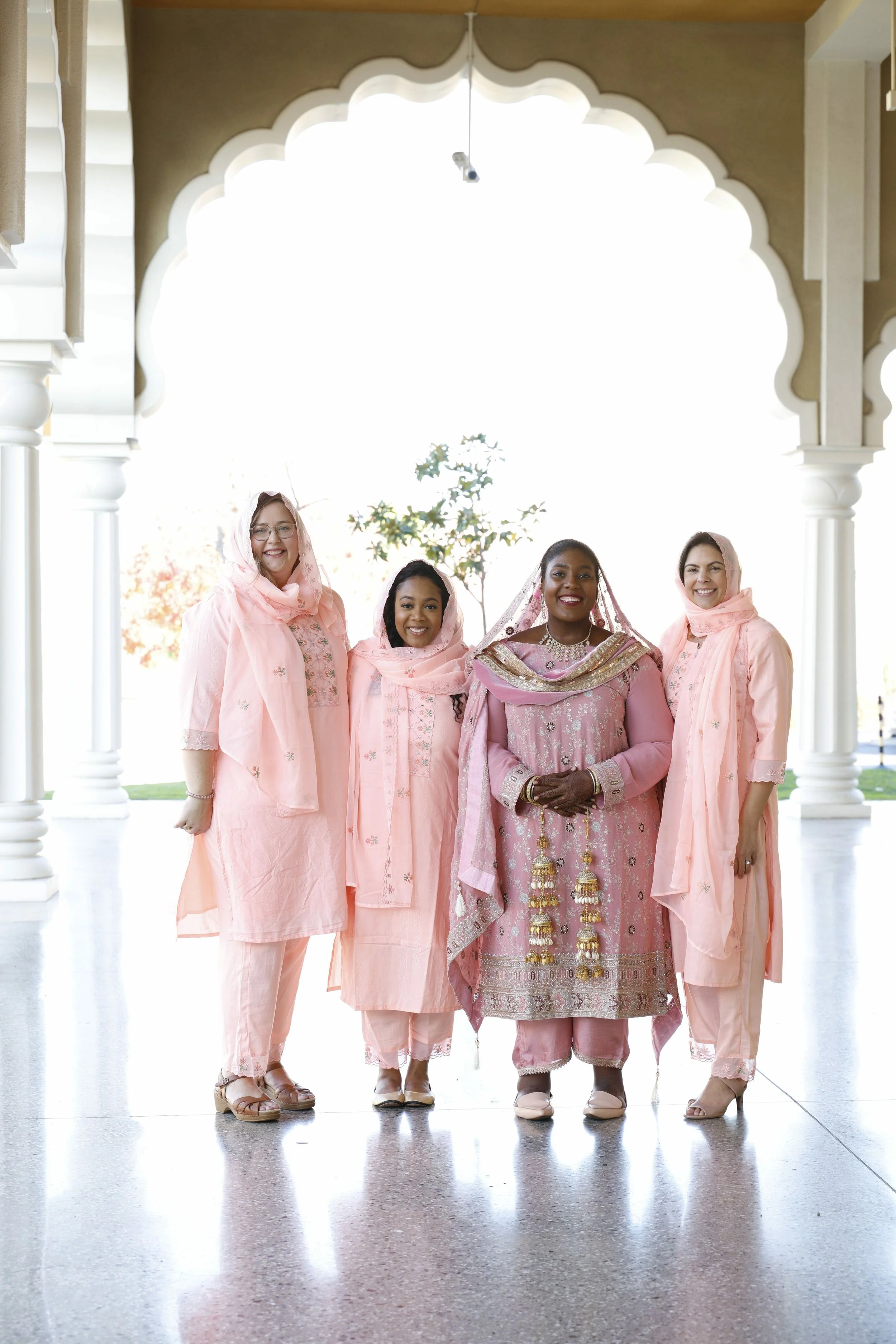 Four women dressed in traditional pink attire, standing under an ornate archway, smiling for a photo at a cultural event.