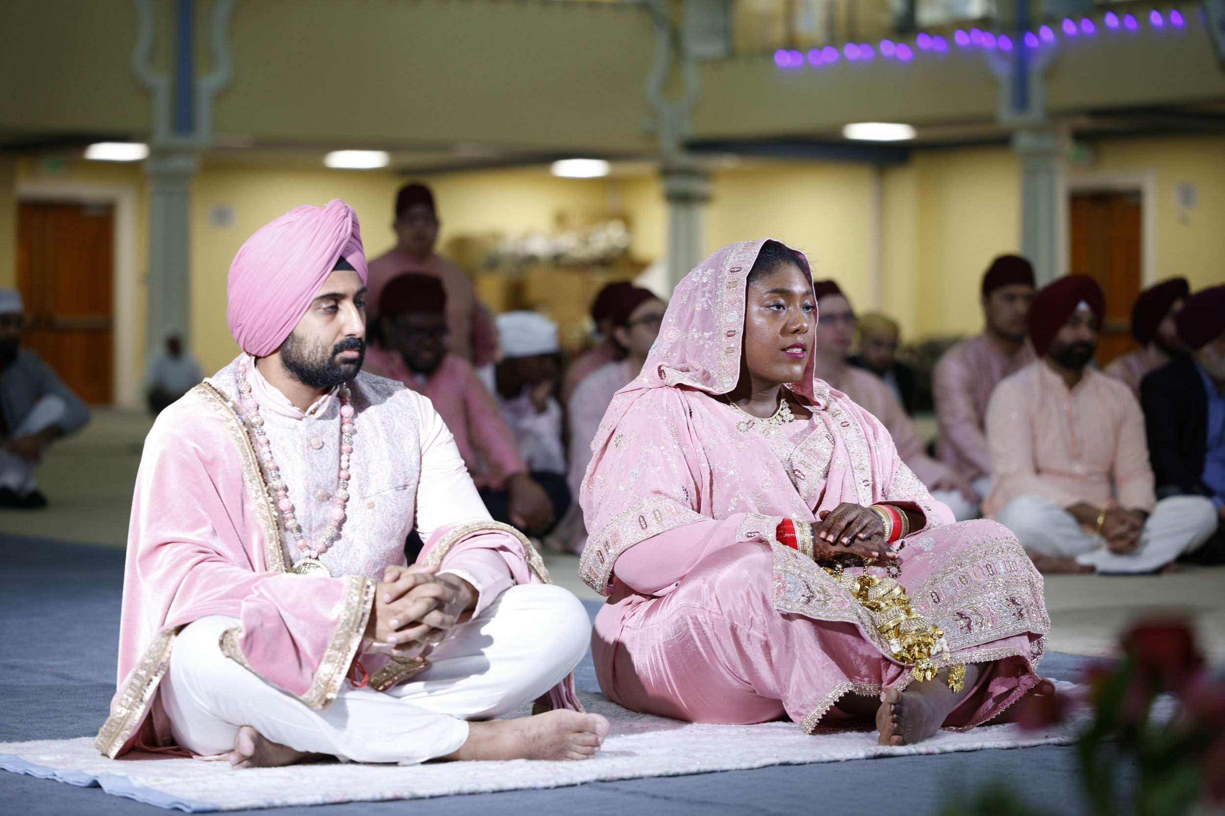 A Sikh couple dressed in traditional wedding attire sitting on the floor during a ceremony, surrounded by guests also dressed in traditional clothing.