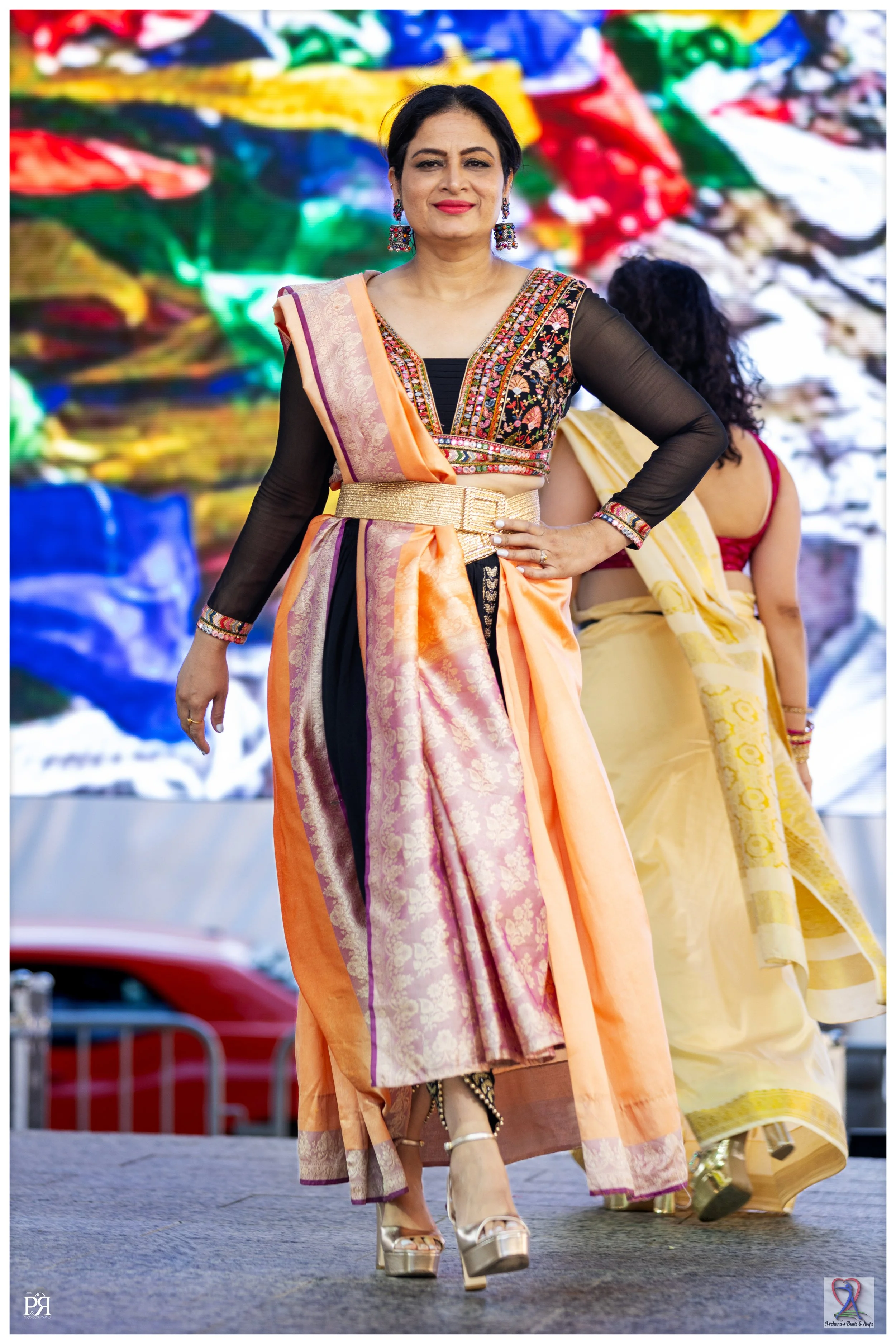 Woman in colorful sari and traditional jewelry walking on stage with a vibrant, abstract background.