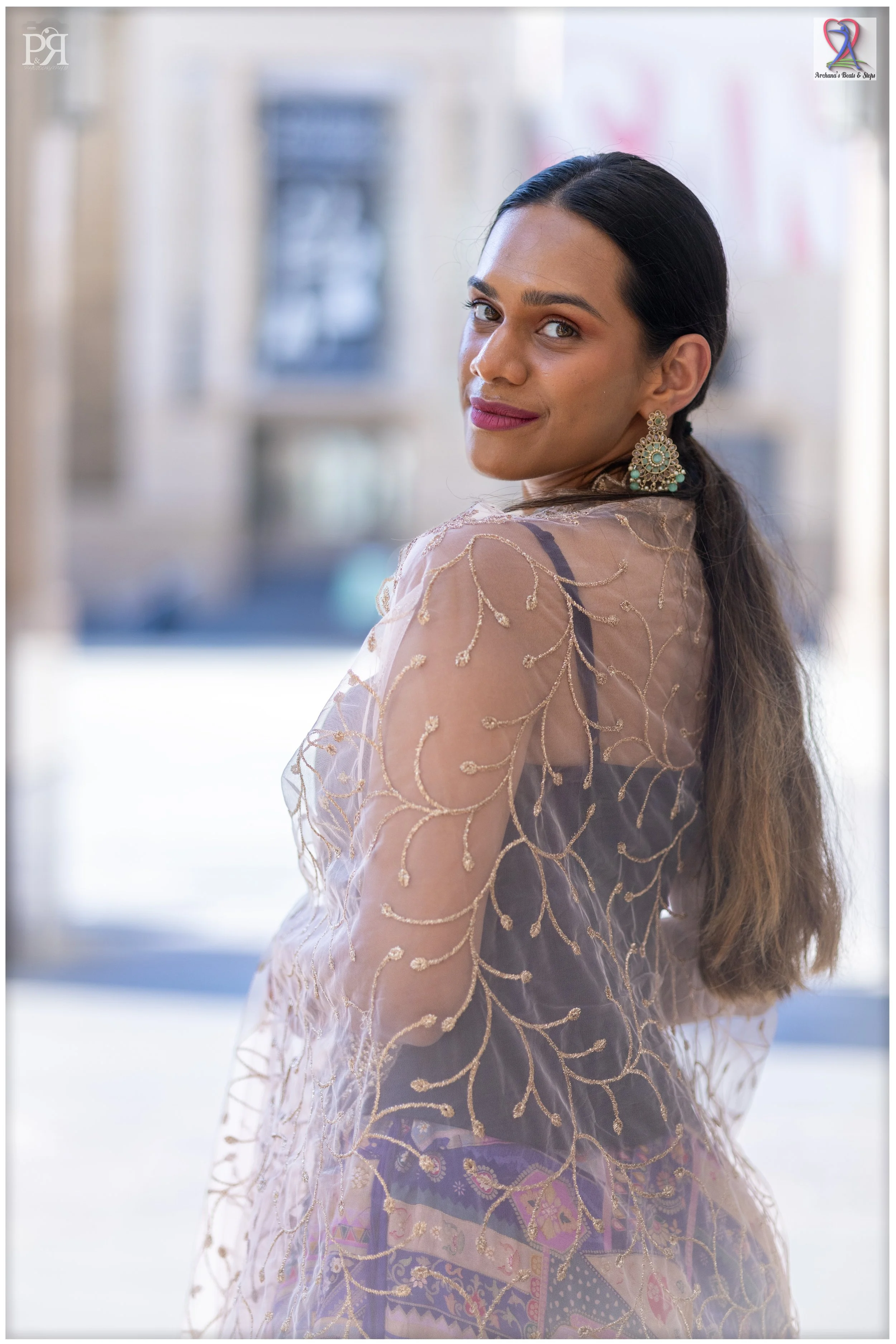 A woman with long dark hair, wearing ornate earrings, in a sheer embroidered dress with gold thread patterns, standing outdoors with a blurred cityscape background.