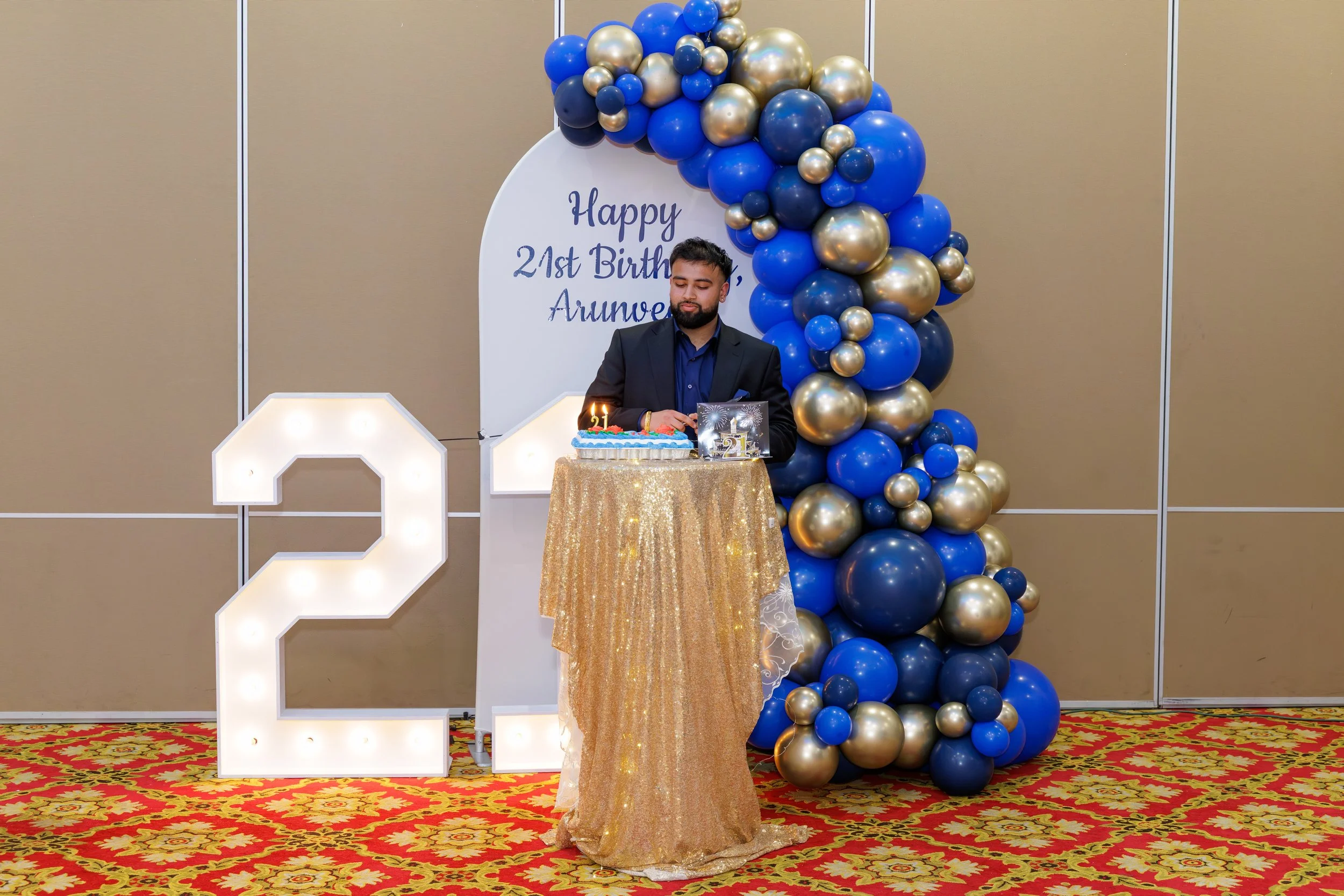 A man in a dark suit celebrating his 21st birthday with a cake, candles, and decorations including a large illuminated number 21 and a balloon arch in blue, gold, and silver, with a sign reading 'Happy 21st Birthday Arunesh' in the background.