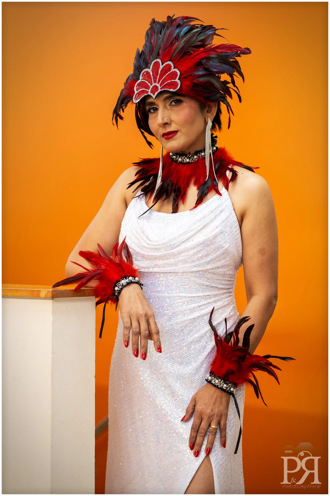 Woman in a white dress and red feathered headdress with matching accessories against an orange background.