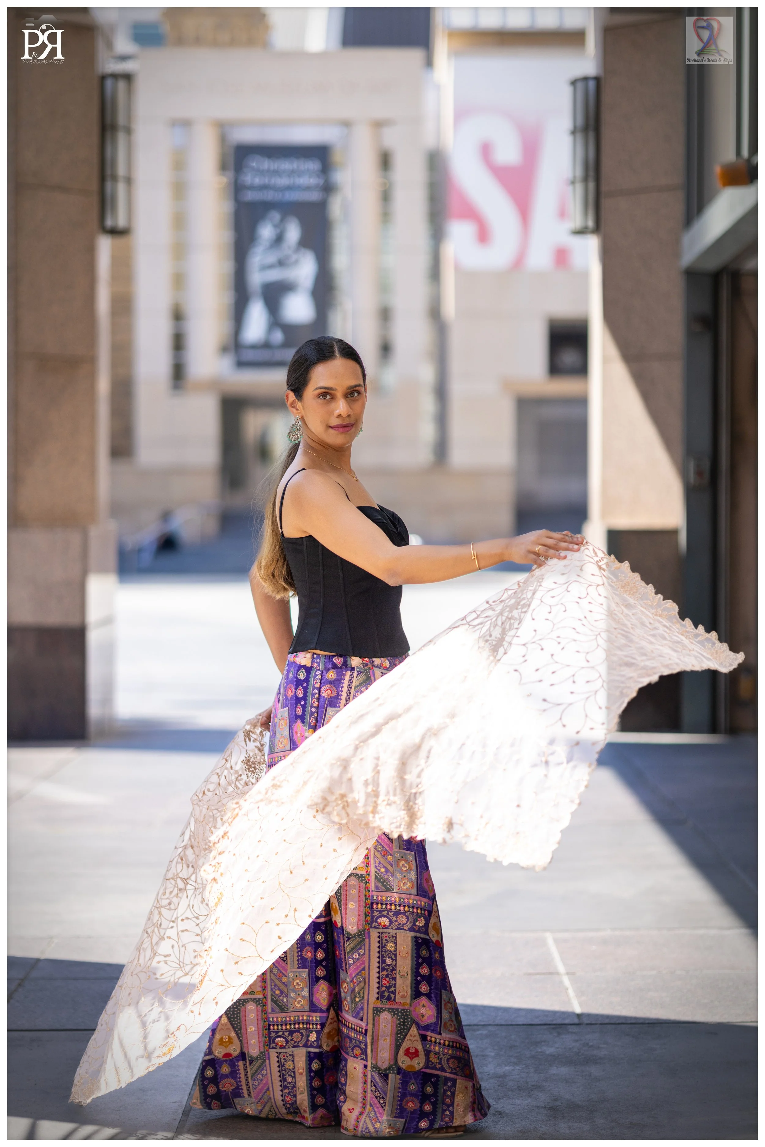 A woman in a black strapless top and colorful patterned wide-leg pants holds a white lace umbrella while standing outdoors in an urban setting, with buildings and a 'For Sale' sign in the background.