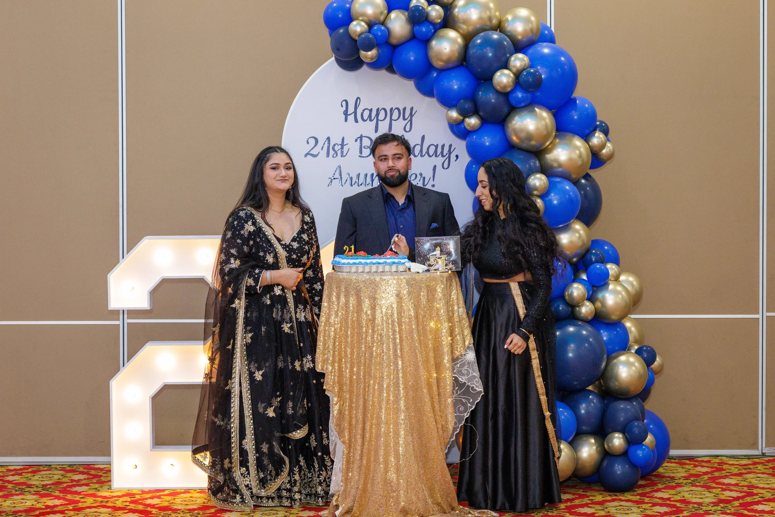 Three people celebrating a birthday with a cake. They are dressed in traditional Indian attire, standing in front of a decorated backdrop with balloons and a sign that says "Happy 21st Birthday, Anumer!"