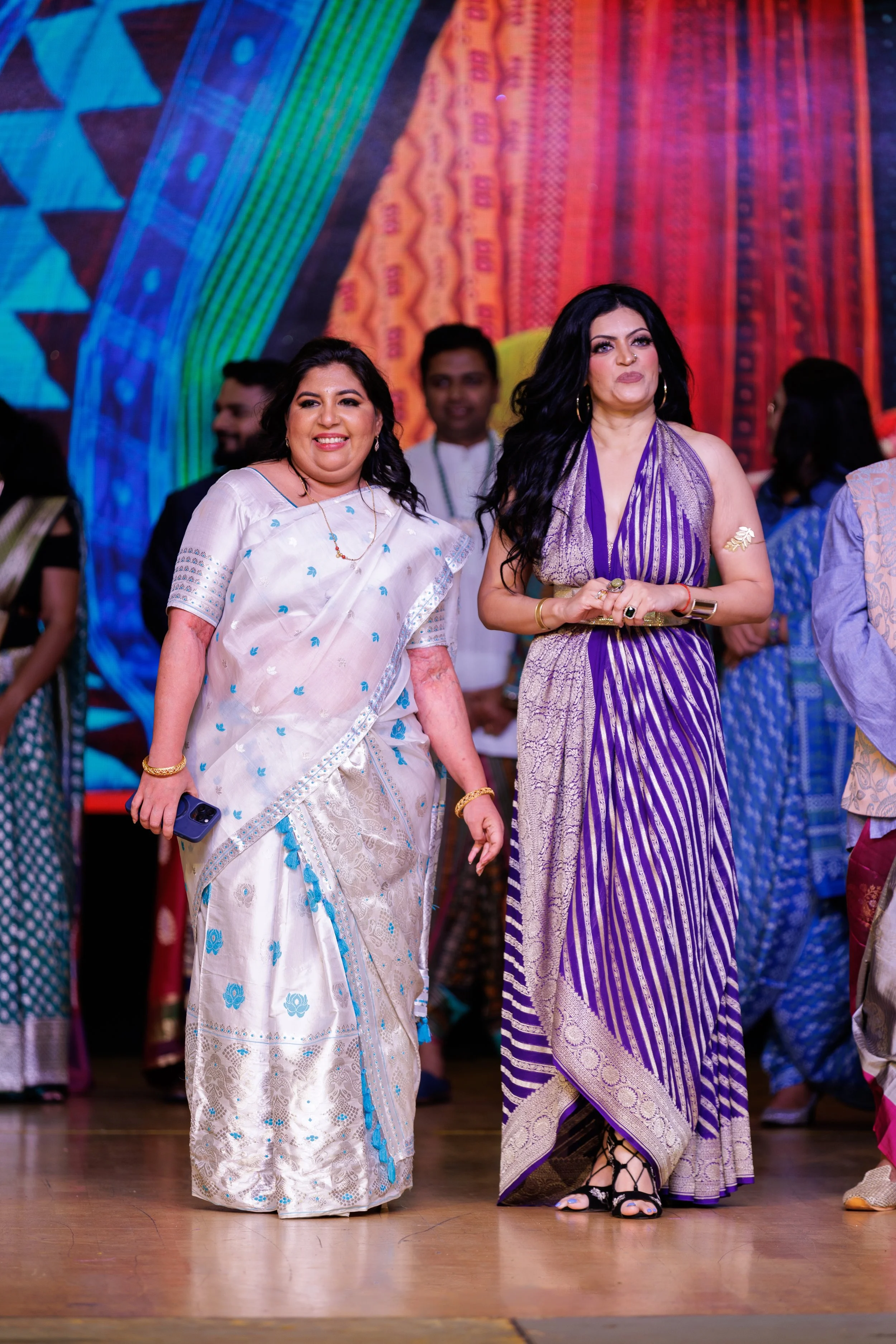Two women dressed in traditional Indian sarees walking on stage at a cultural event, with colorful decorations and other attendees in the background.
