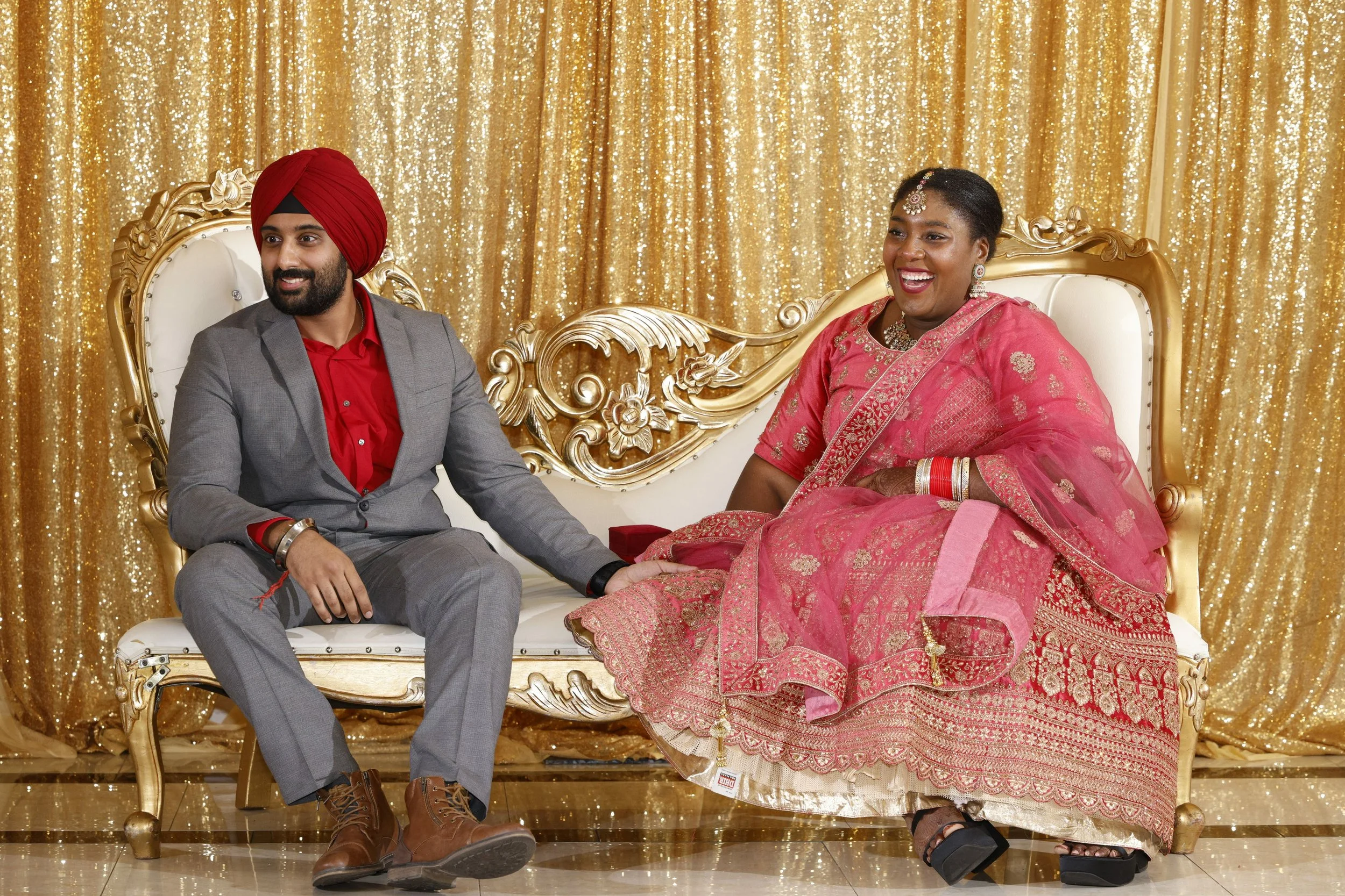 A man in a gray suit and red shirt sitting on a white and gold ornate sofa, holding hands with a woman in a pink traditional dress, sitting on a matching sofa with gold details, against a gold sequin backdrop, both smiling.
