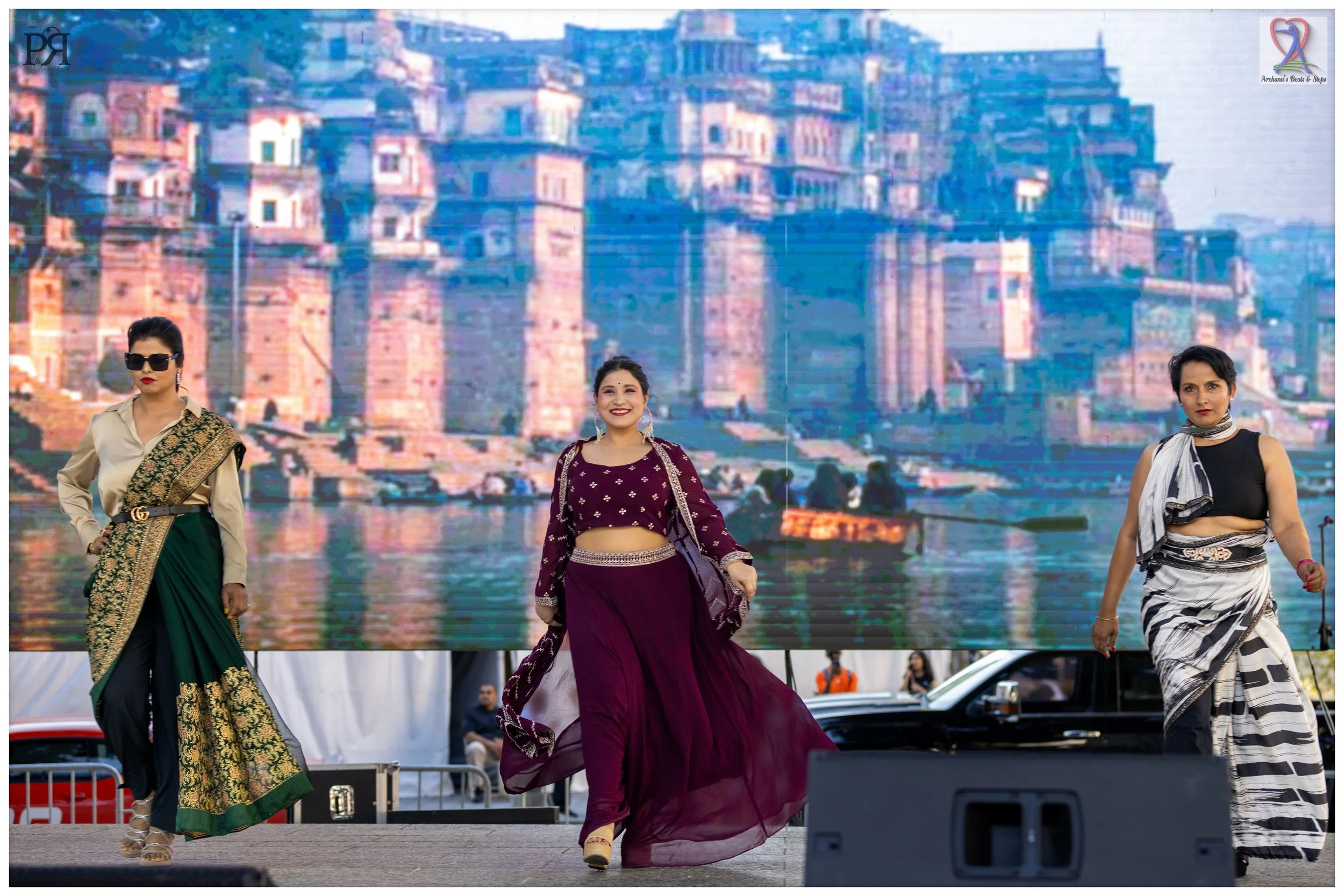 Three women walk on stage during a cultural dance event, with a large digital screen displaying a cityscape of colorful buildings behind them.