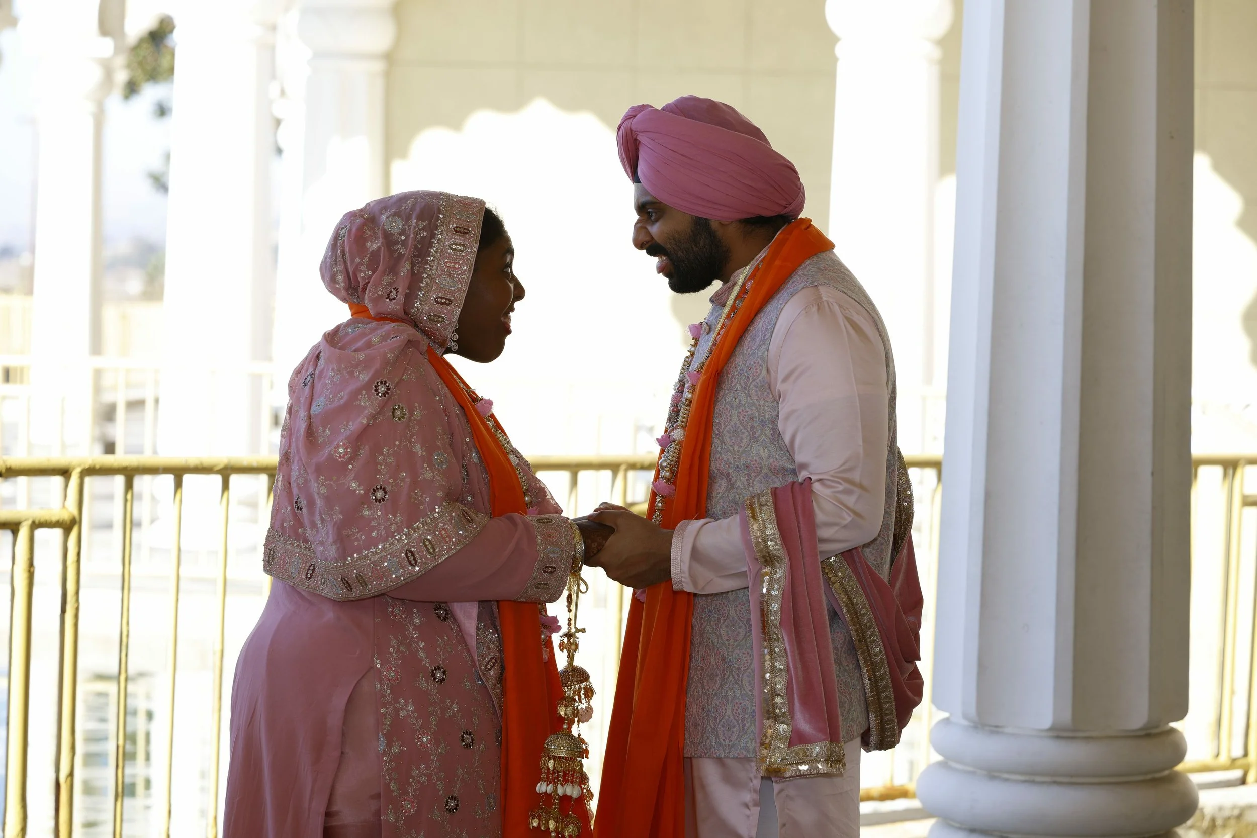 A couple dressed in traditional Indian wedding attire standing face-to-face and holding hands on a balcony with white columns.