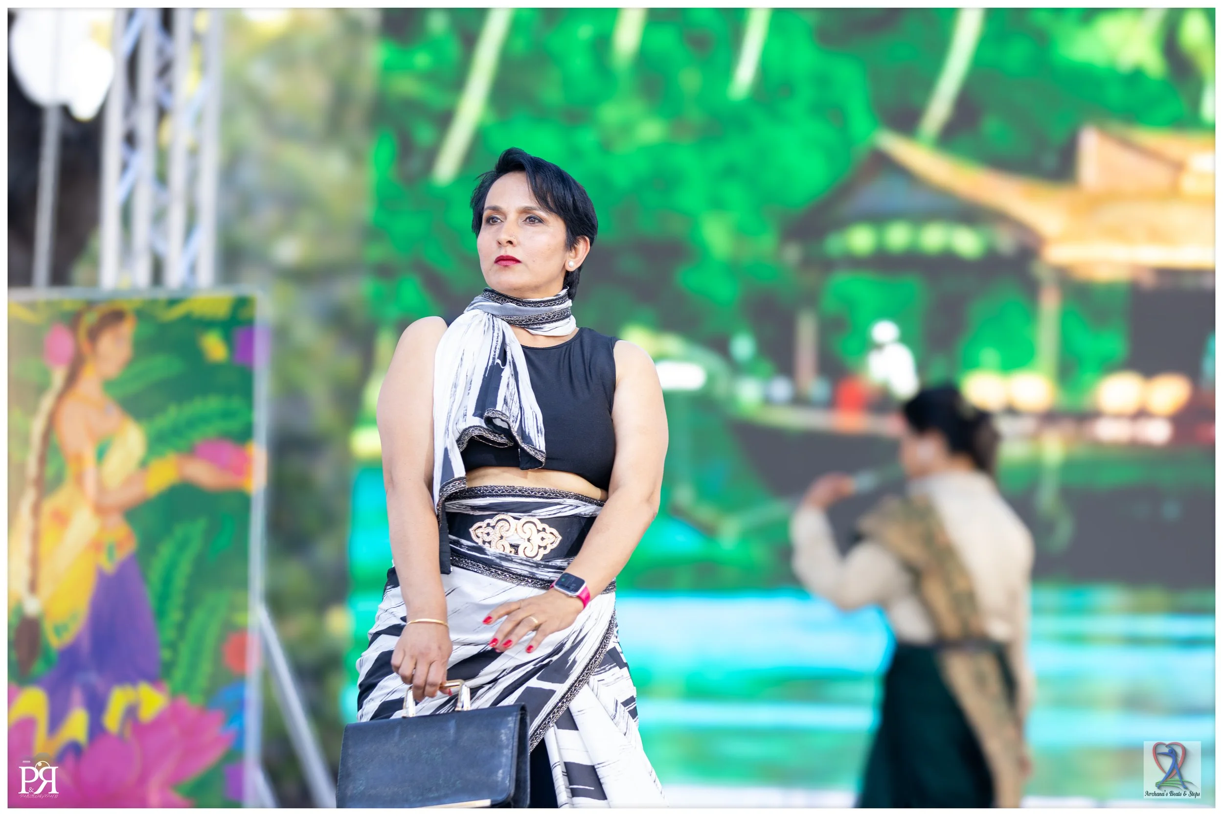 A woman with short black hair wearing a black sleeveless top and a traditional black and white wrap skirt, holding a black handbag and wearing a smartwatch, standing outdoors with a vibrant, colorful green background and a blurred woman in traditiona